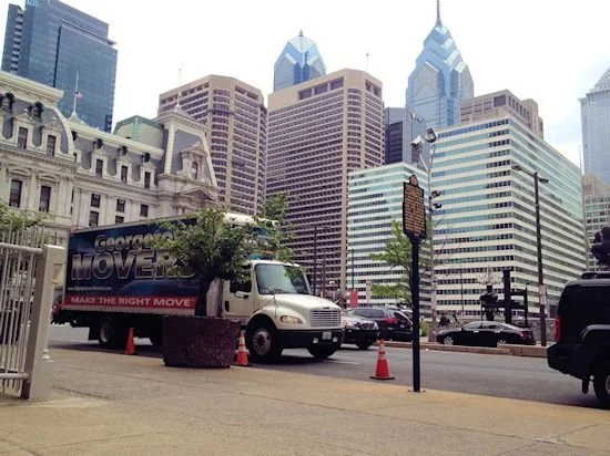 Street view in a downtown area with tall skyscrapers, a moving truck labeled "George's Movers," traffic cones, parked cars, and a few trees.