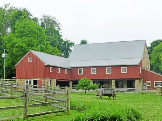 Red barn with a gray metal roof, surrounded by green trees and grass, with a wooden fence and a horse in the foreground.