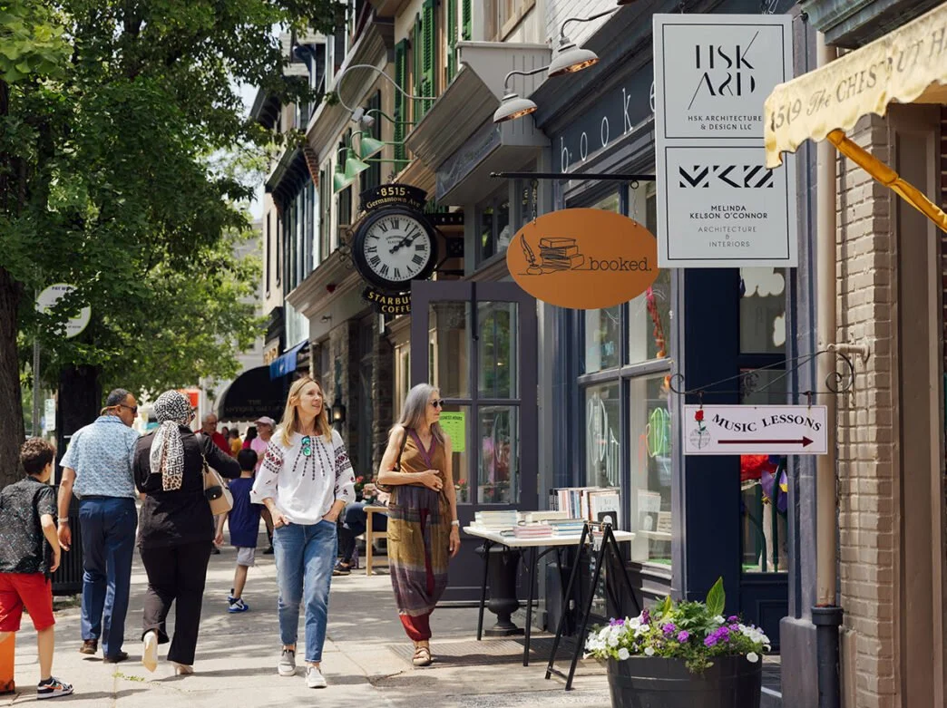People walking on a city sidewalk lined with shops, including a bookstore with signs for architecture, interiors, and music lessons, a Starbucks coffee shop, and other stores, with trees and a clock displaying 2:10.