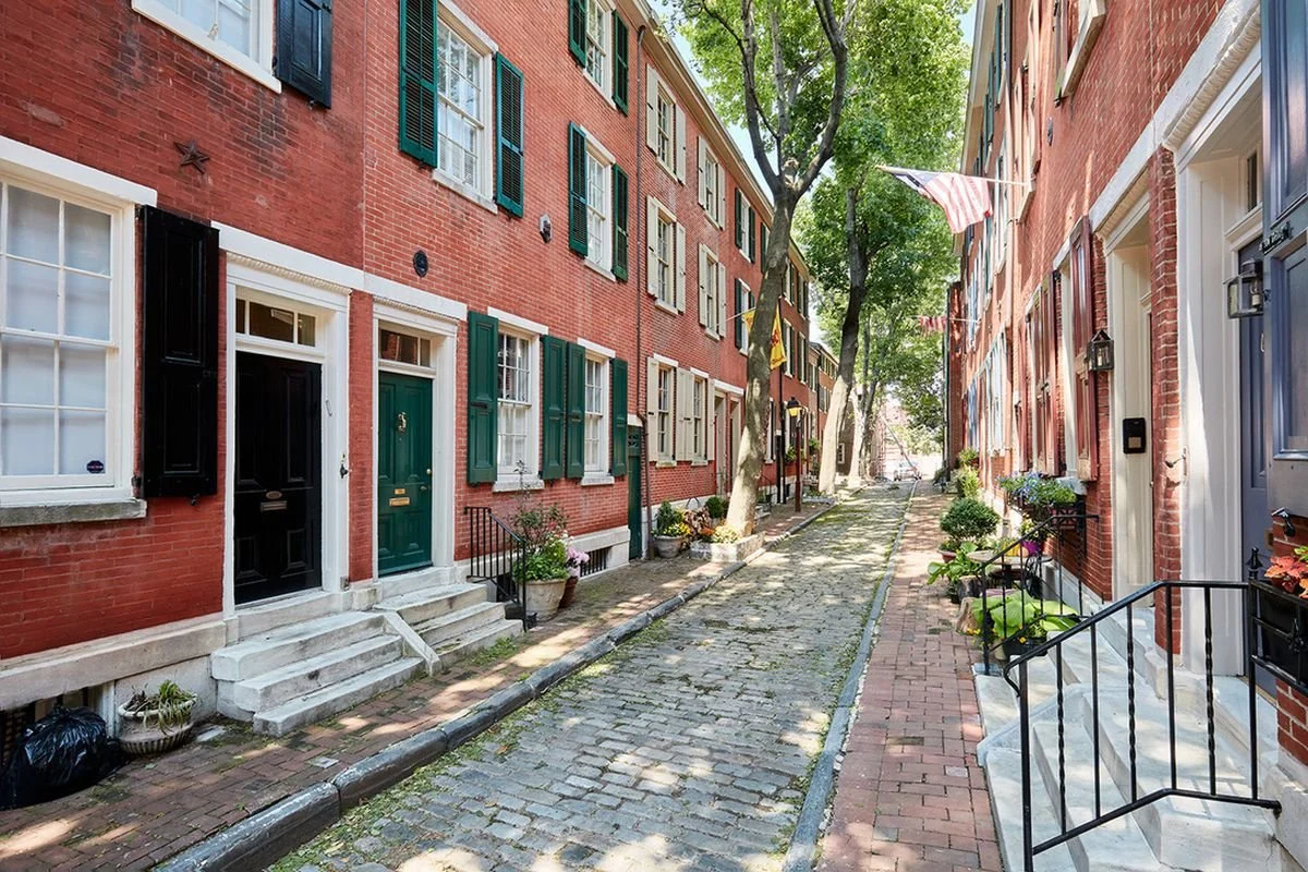 A narrow cobblestone street lined with brick townhouses, some with green shutters and American flags, trees providing shade, and potted plants outside the doors.