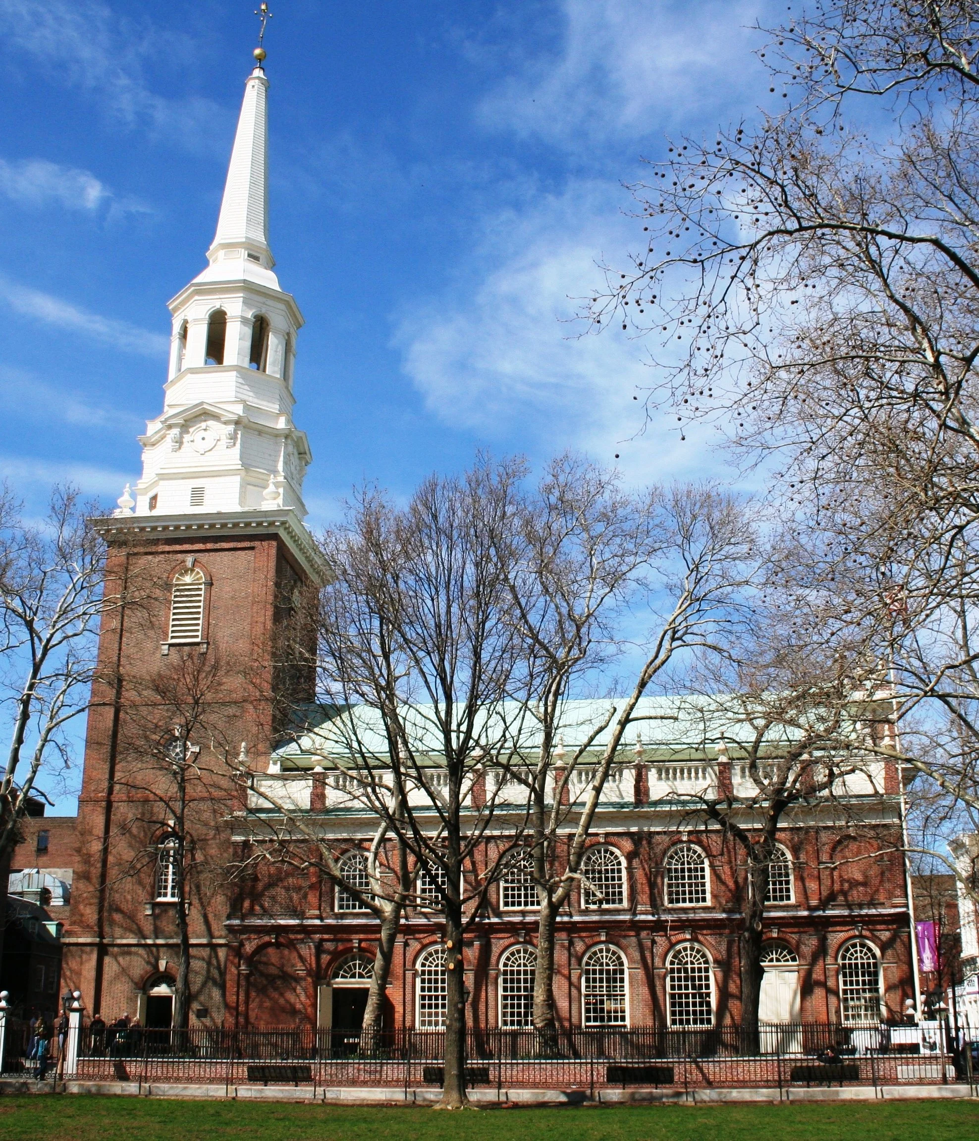 Historic brick church with white steeple and arched windows, trees in front, blue sky with some clouds.