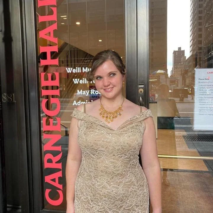 A young woman in a beige lace dress and gold jewelry standing in front of a glass door with red text, smiling at the camera.