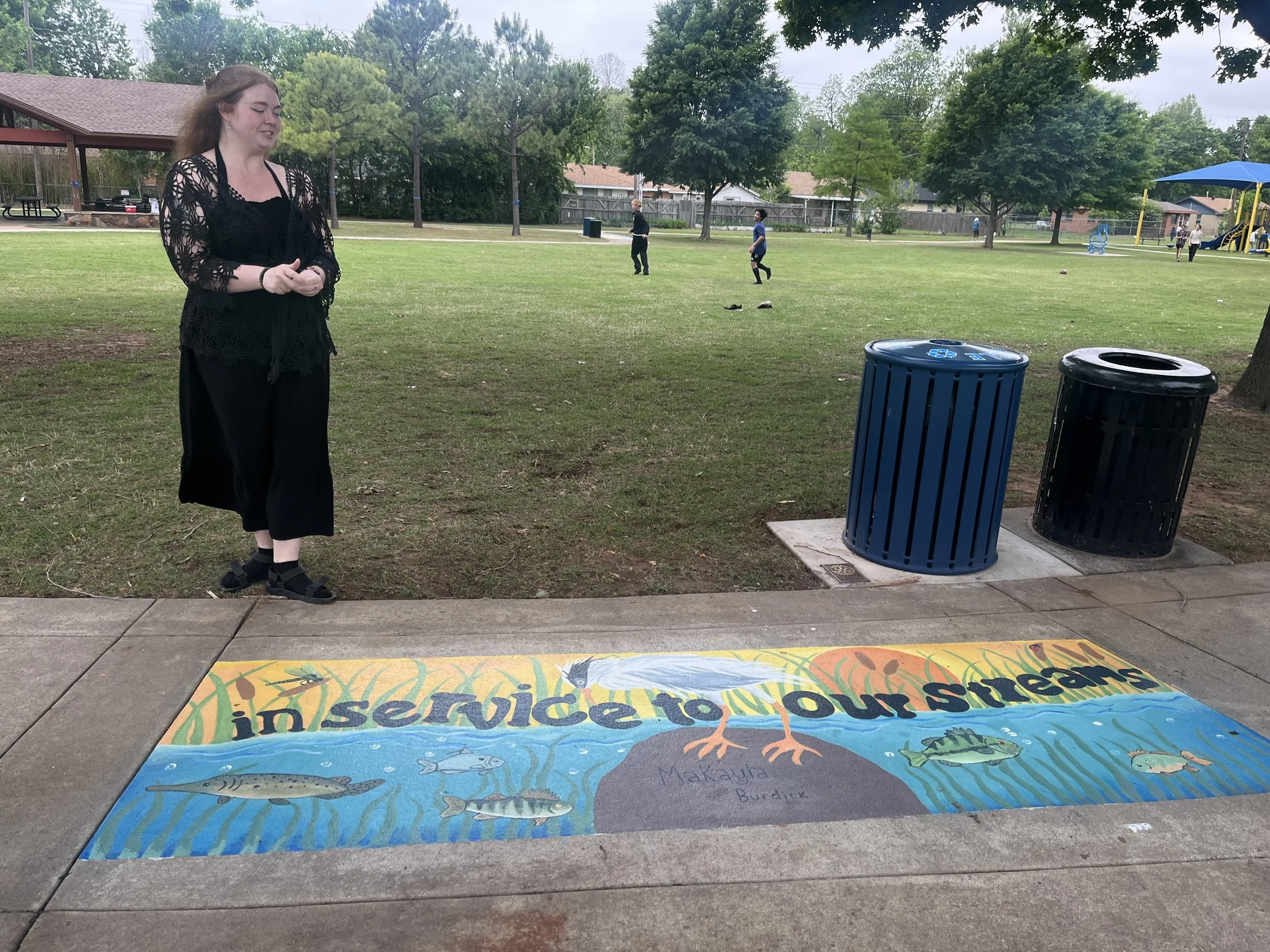 Artist Makayla Burdick with their Artful Inlets mural on the sidewalk at Rotary Park.