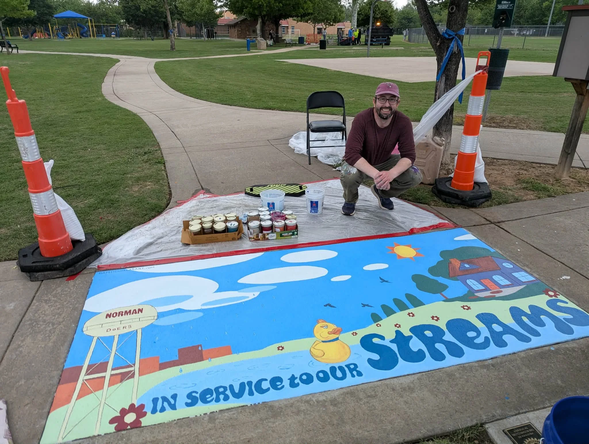 Artist Sam Douglas with their Artful Inlets mural on the sidewalk at Rotary Park.