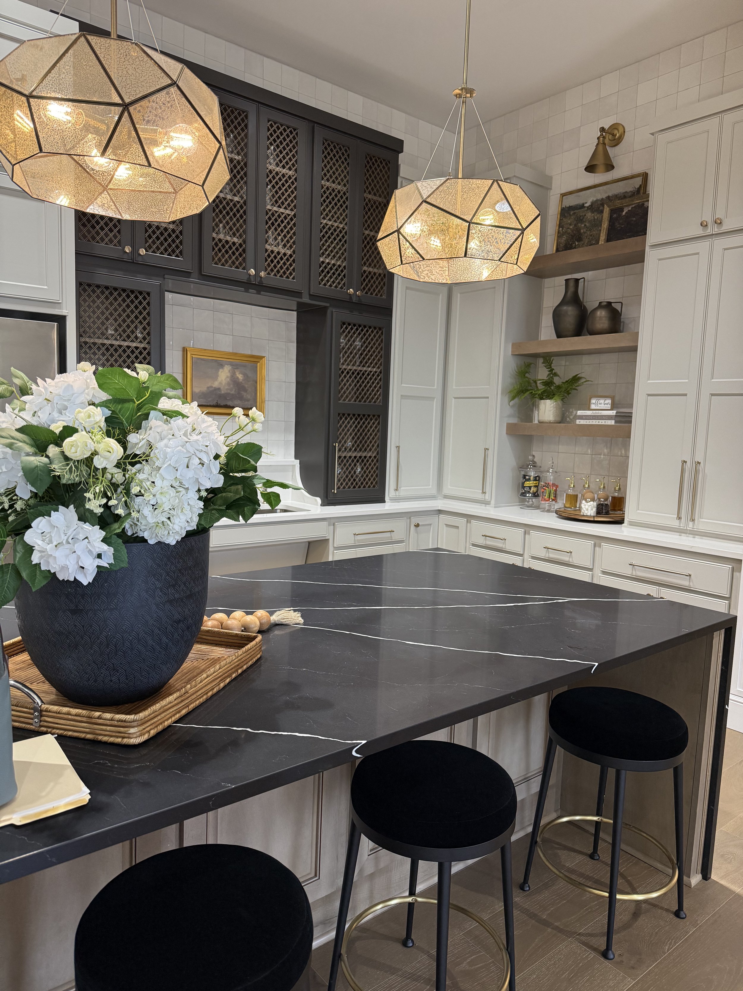 Modern kitchen with black and white cabinetry, a black marble island with white veining, black bar stools, and decorative shelves with vases and plants.