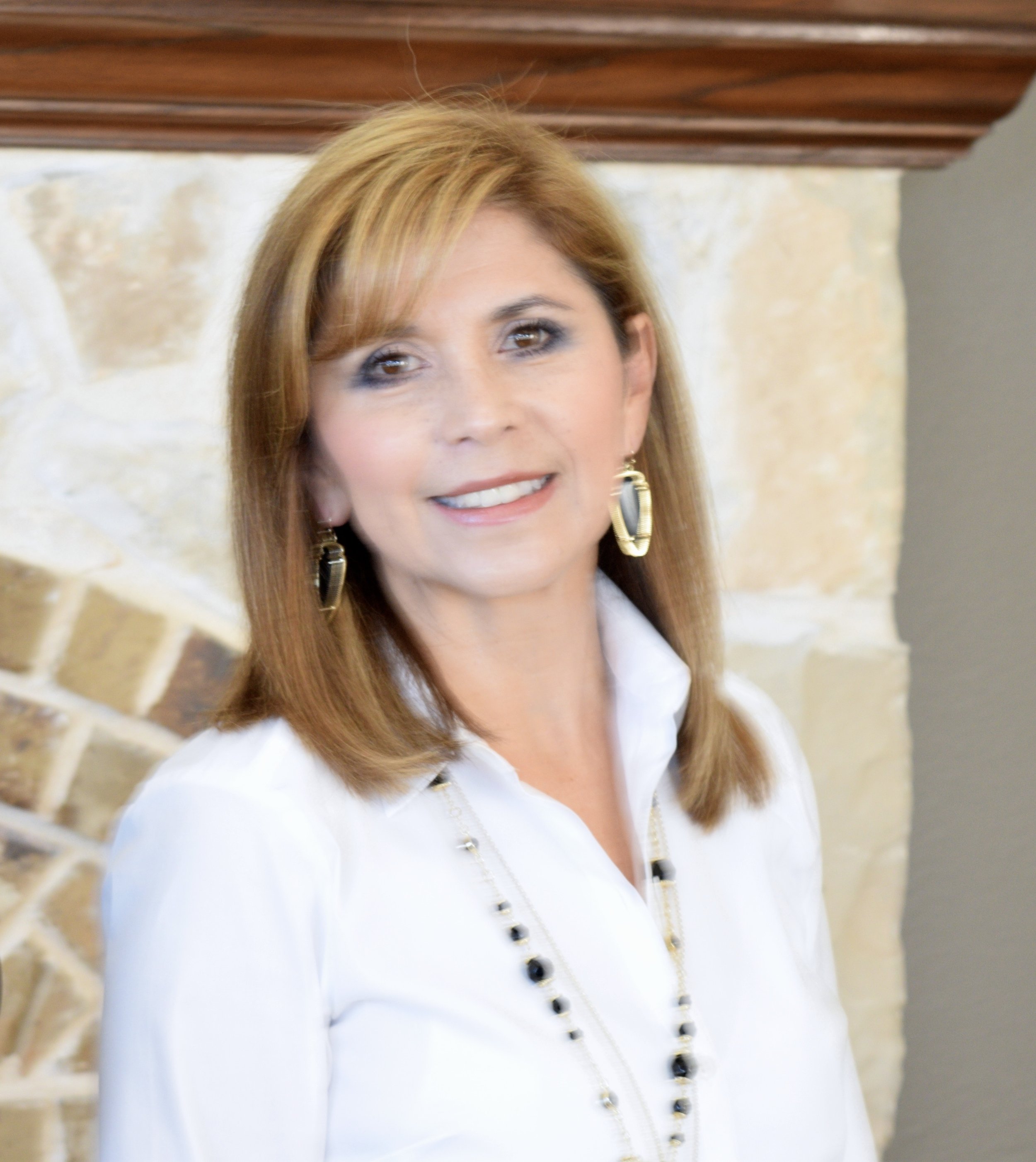 A woman with shoulder-length light brown hair, wearing a white blouse, black and gold earrings, and a black and gold necklace, standing in front of a stone wall with a wooden mantle above.