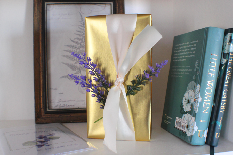 Decorative golden gift box with white ribbon and lavender sprigs, positioned between a framed botanical print and three teal-colored books on a white shelf.