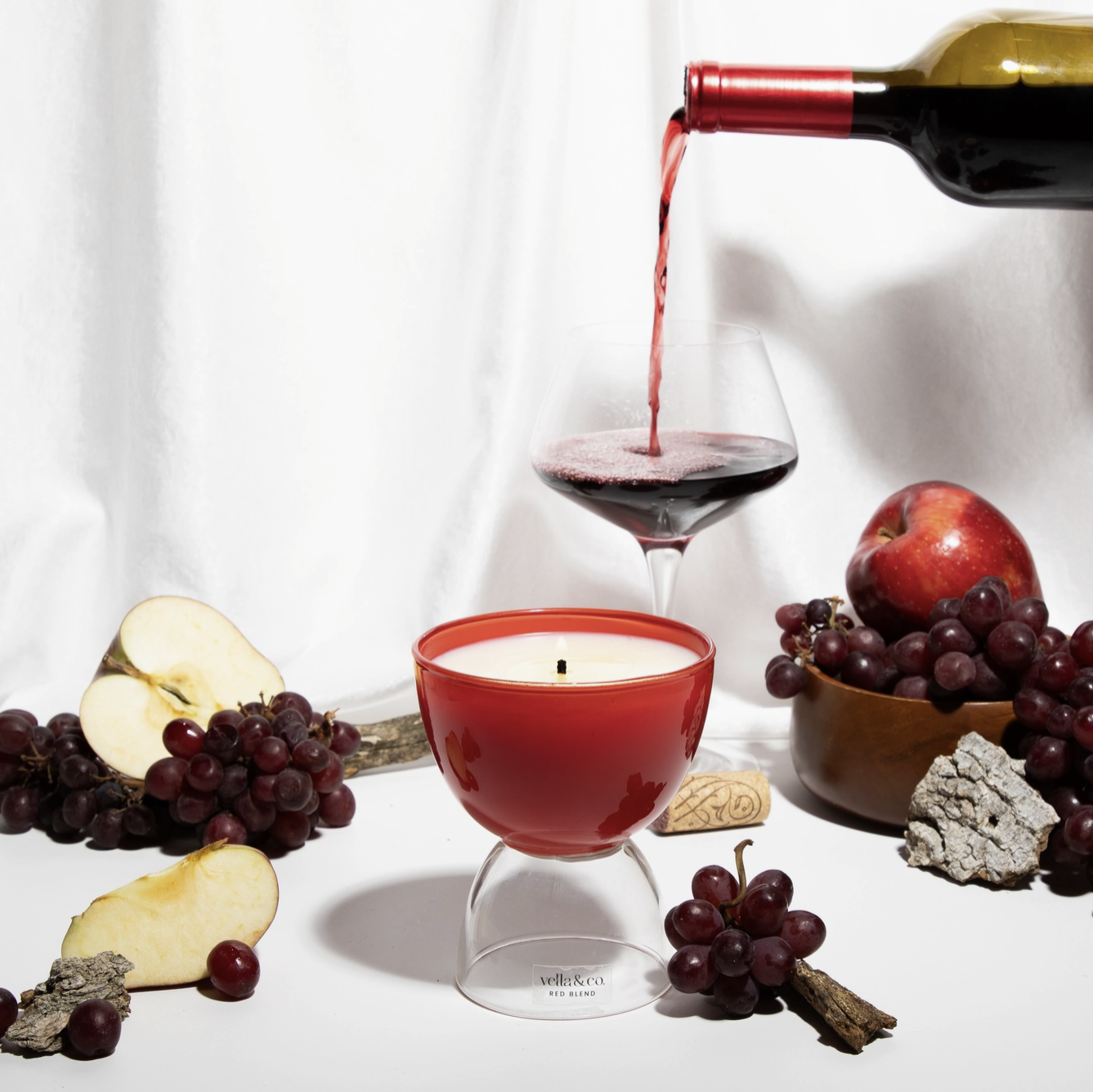 A bottle of red wine being poured into a glass, with grapes, apple slices, an apple, a candle, and decorative stones on a white background.