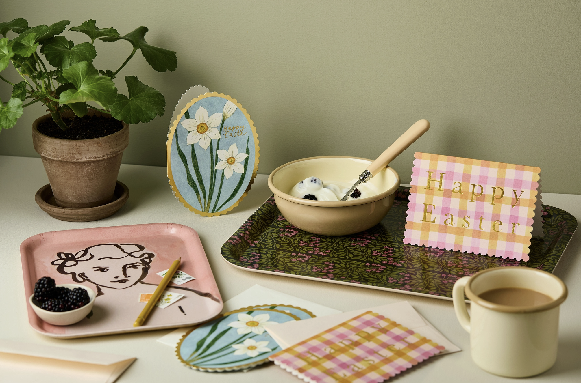 Easter greeting card with the words "Happy Easter" in gold letters, placed on a decorative tray with a bowl of yogurt and blueberries, a cup of coffee, a potted plant, and Easter-themed illustrations and decor on a table.