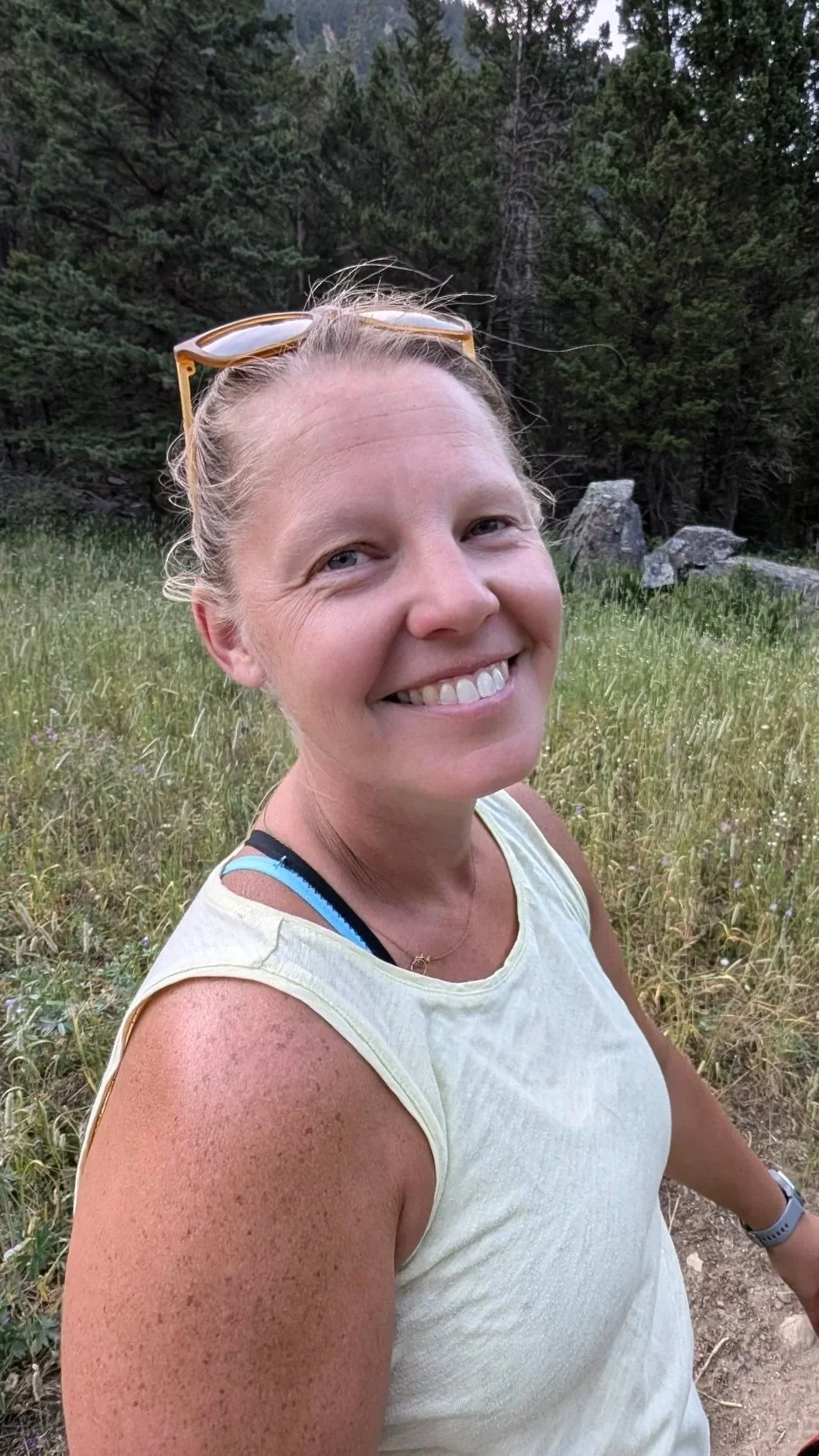 A woman with long brown hair, wearing a green shirt, smiling outdoors with trees and grass in the background.