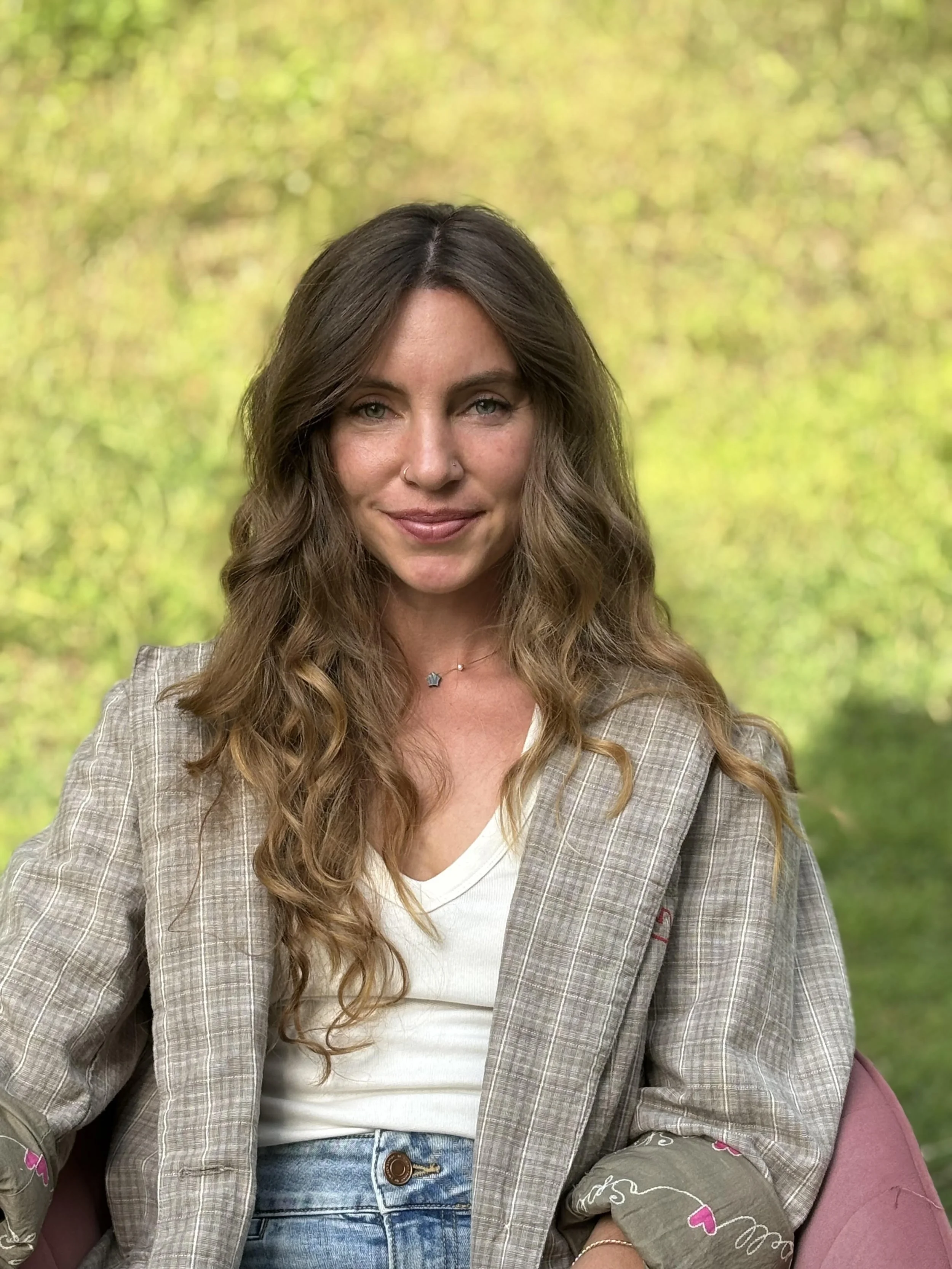 A woman with long wavy brown hair and blue eyes sitting outdoors in front of a blurred green background, smiling slightly, wearing a checked blazer over a white shirt and blue jeans.
