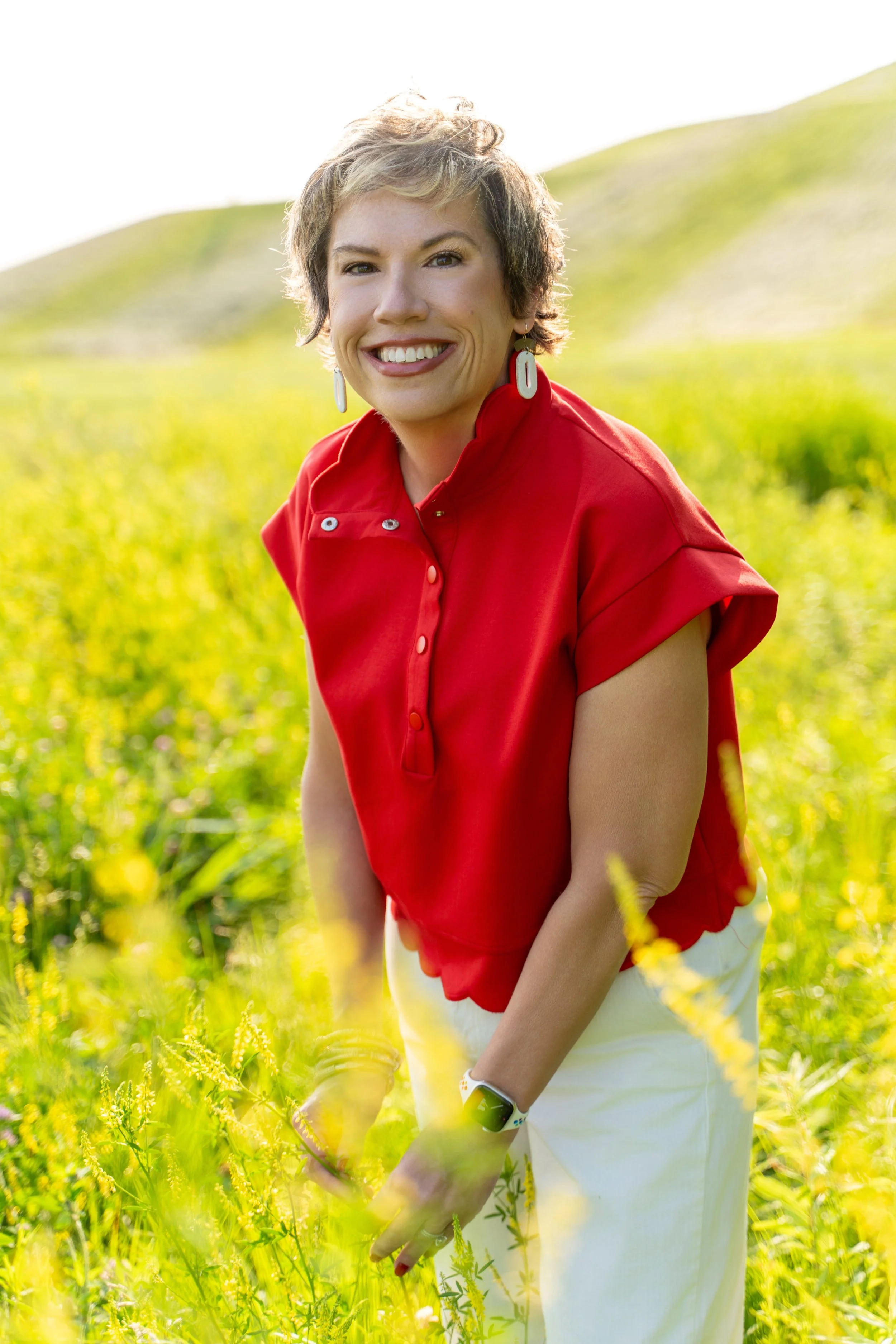 Woman with short blondish-brown hair smiling, wearing a red top and earrings, standing in a yellow flower field with hills in the background.
