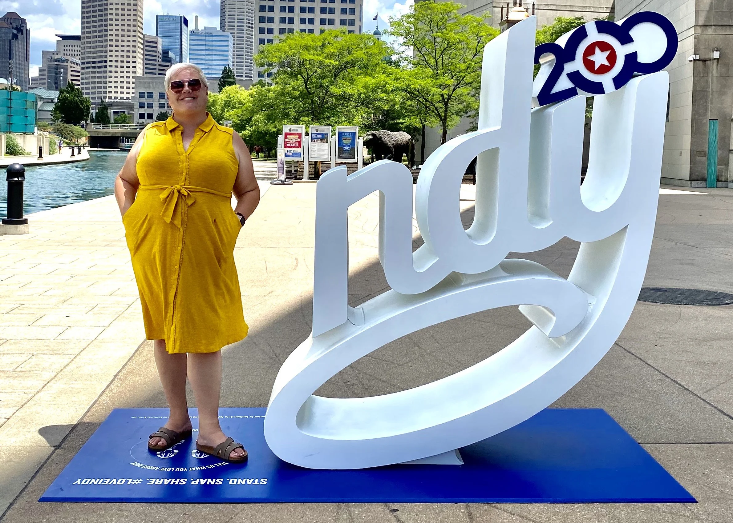 A woman in a yellow dress and sunglasses standing next to a large white Indy sign with a red star inside the zero, on a blue platform that reads 'Stand, Snap, Share, Love Indy'.