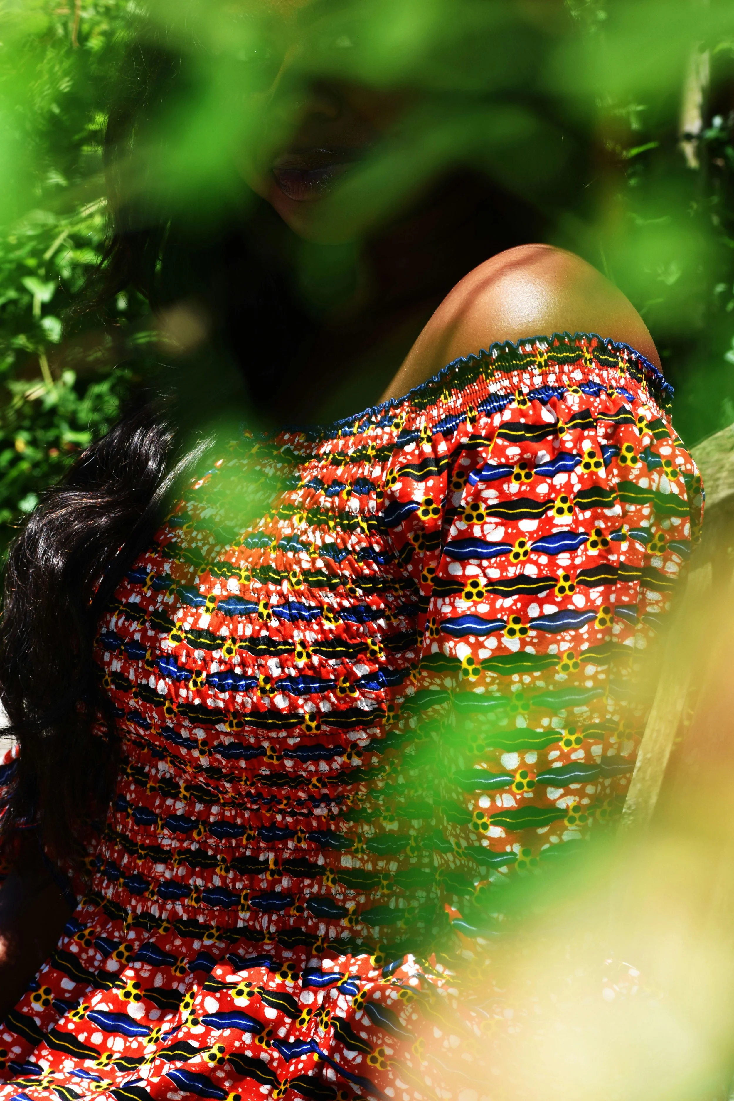 Person with long dark hair wearing a colorful patterned off-shoulder top, partially obscured by green leaves.