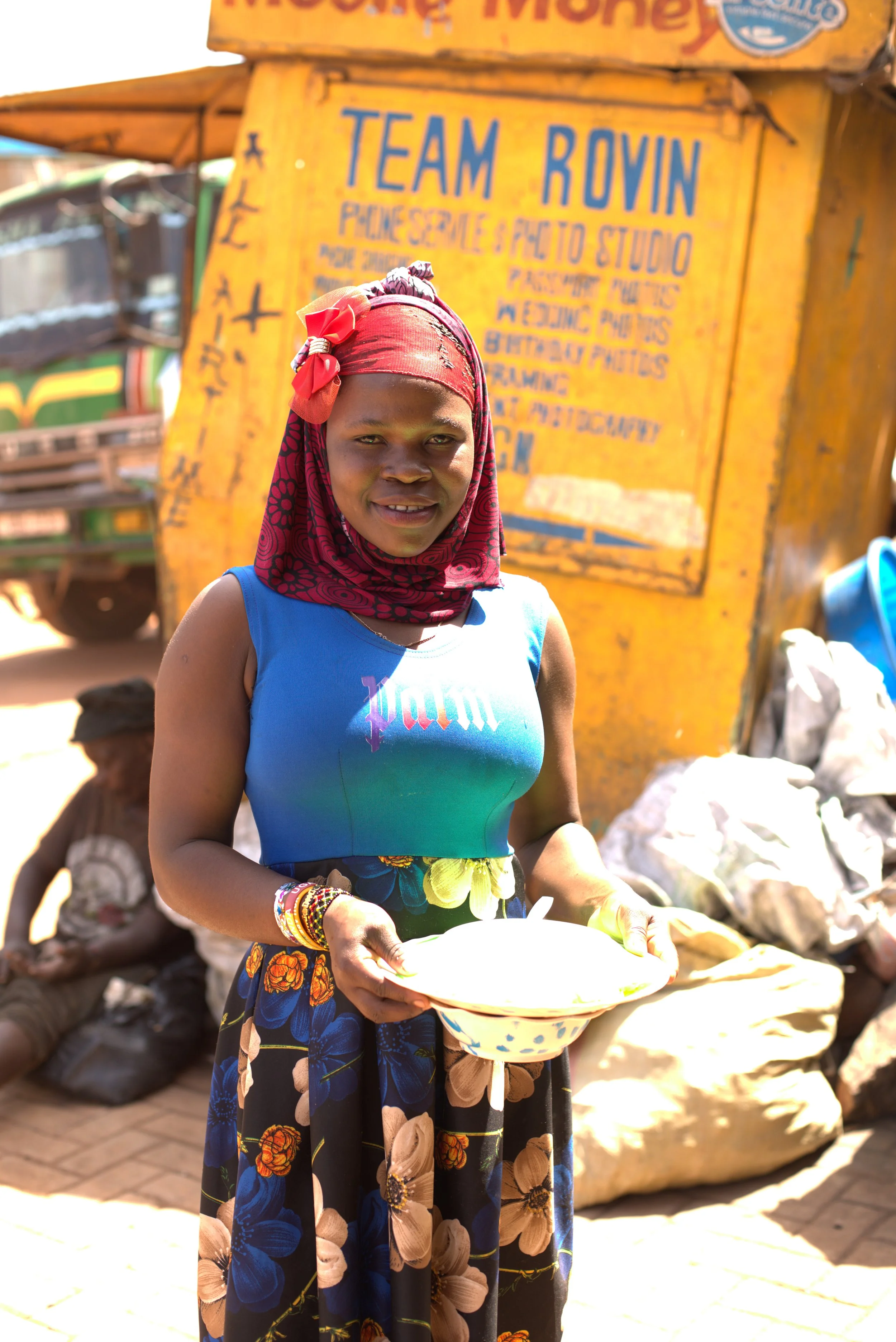 A woman standing outdoors holding a white bowl with a blue dot pattern, wearing a red headscarf, a blue sleeveless top, and a floral skirt. There are people and a yellow sign in the background.