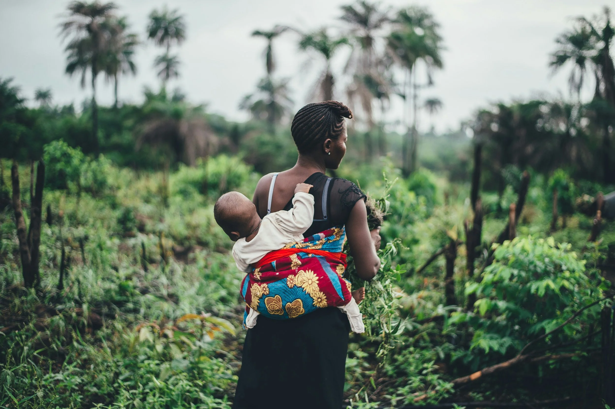 A woman carrying a sleeping child on her back in a green, rural area with palm trees in the background.
