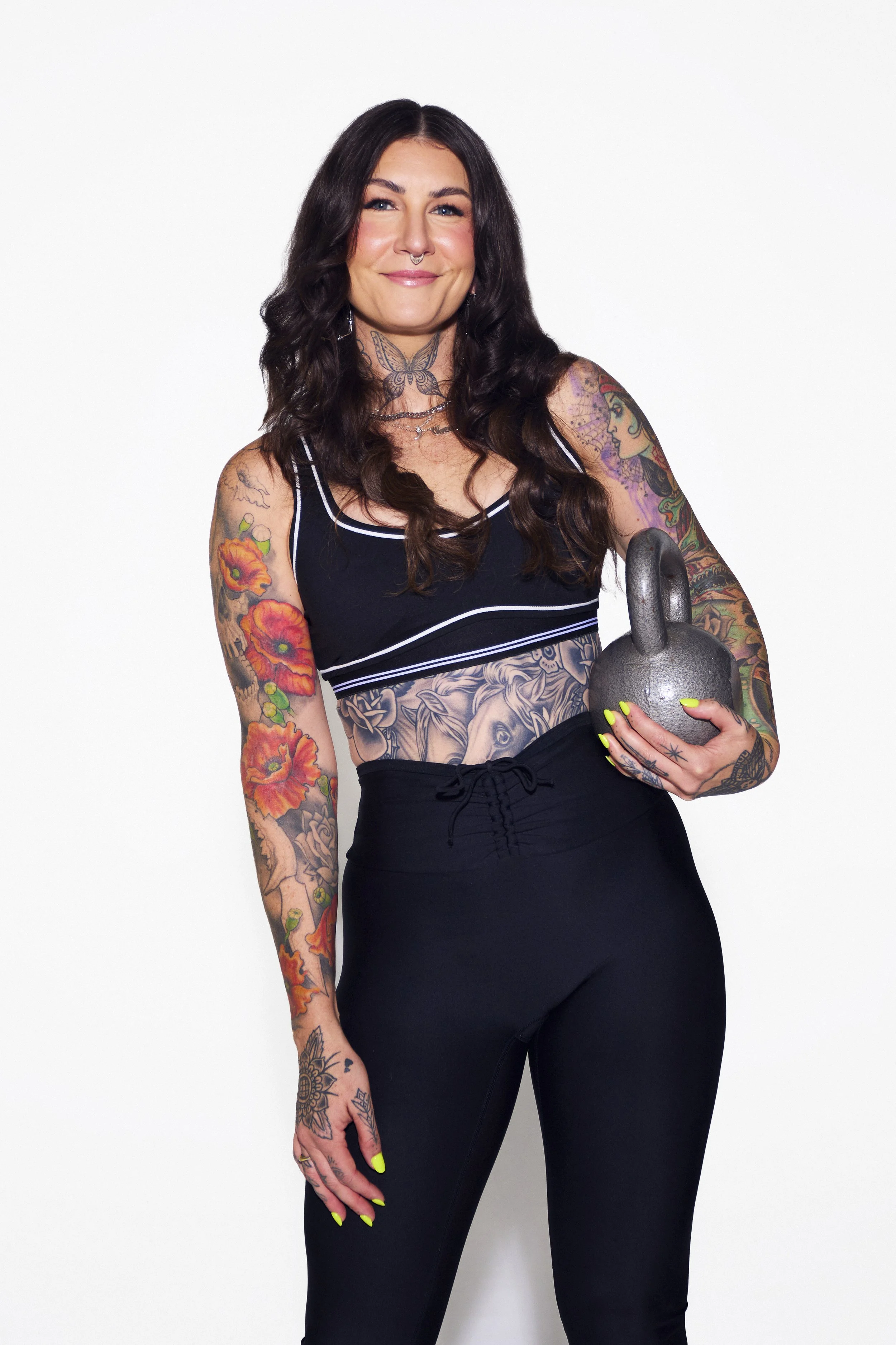 A woman with long dark hair, tattoos on her arms and torso, wearing a black workout tank top and black workout pants, holding a kettlebell, and standing against a plain white background.