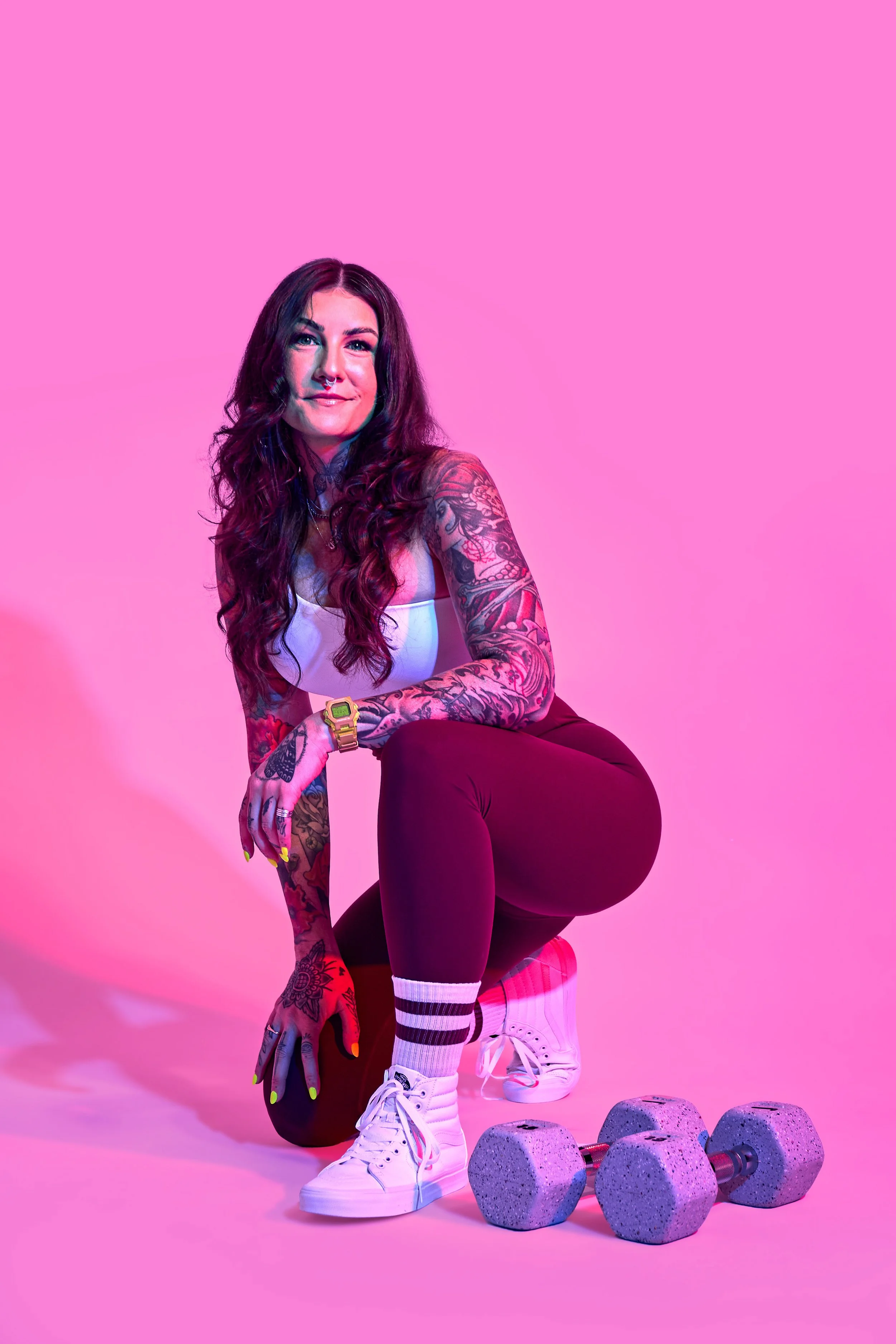 A woman with tattoos, long dark hair, and bright makeup kneeling on one knee in front of pink background with four gray dumbbells, wearing white sneakers, striped socks, black leggings, and a white tank top, smiling at the camera.