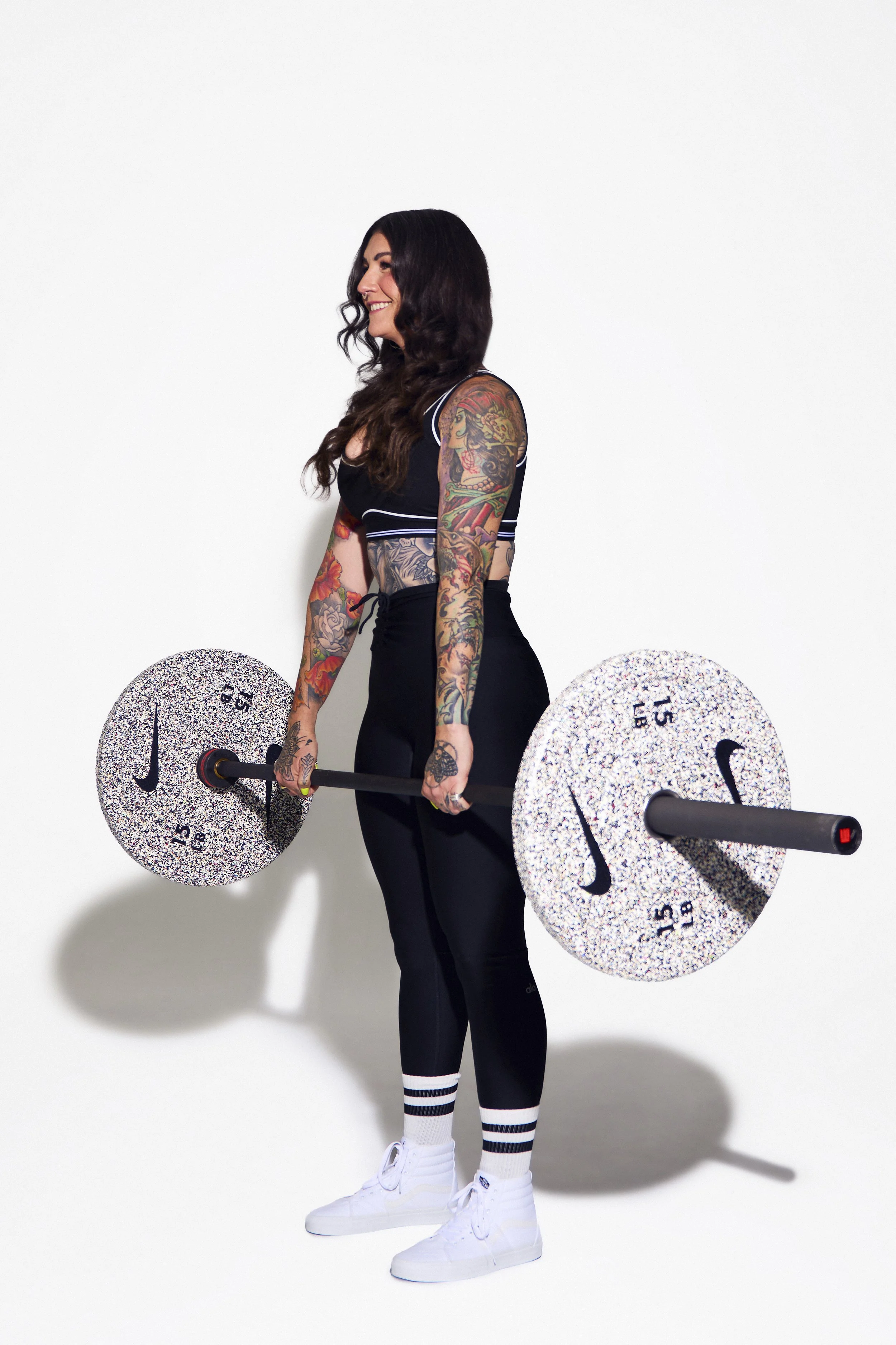A woman with tattoos standing against a white background, holding a barbell with colorful, speckled weight plates.