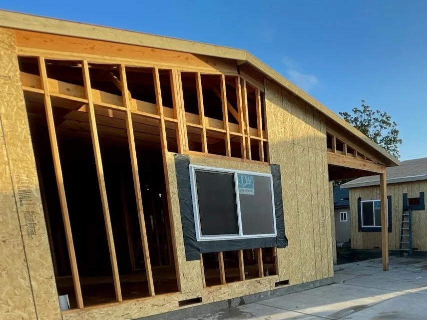 Front view of a house under construction with exposed wooden framing, partially installed window, and plywood sheathing, with a clear blue sky in the background.