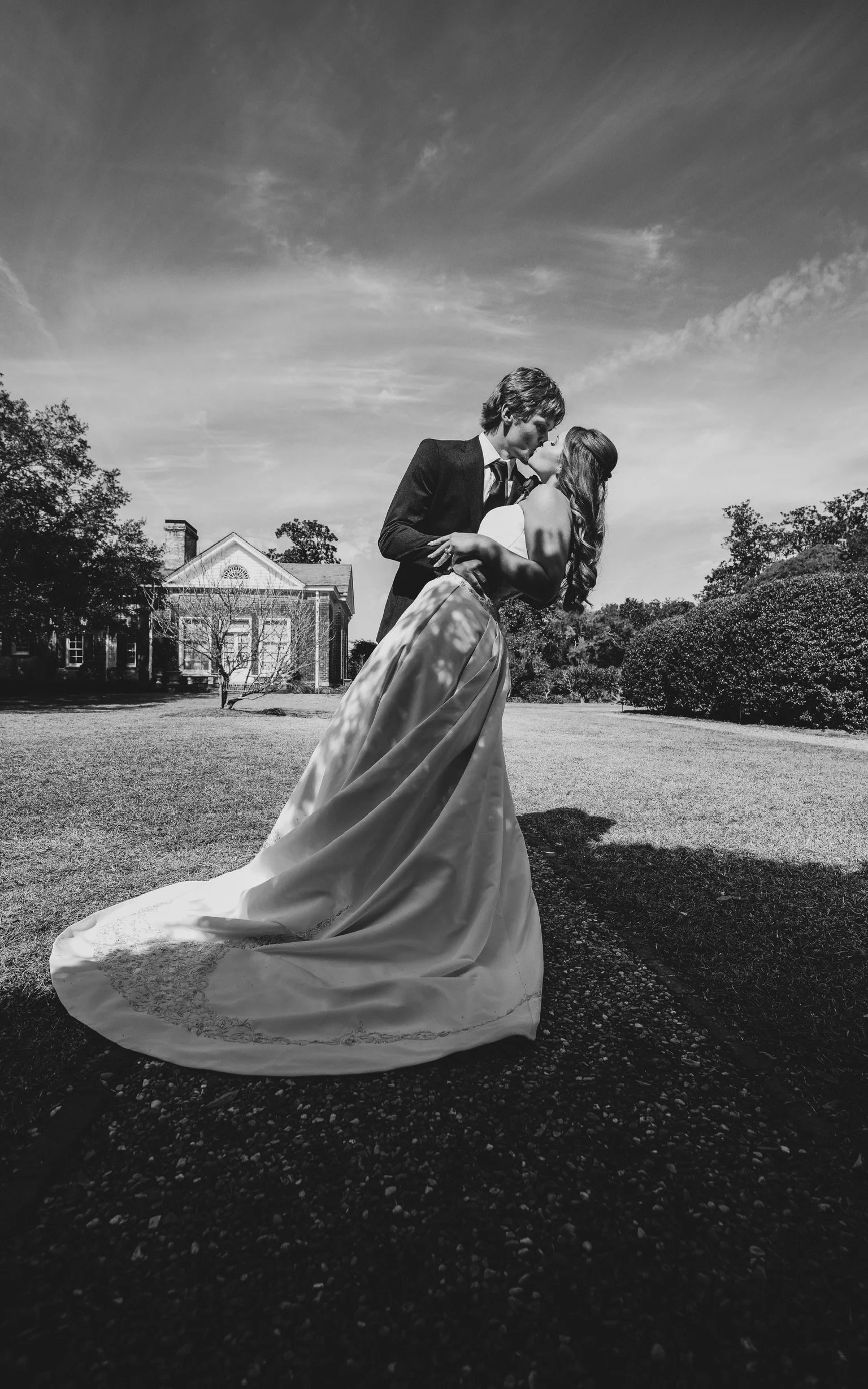 A black and white photo of a bride and groom kissing outdoors, with a large house and trees in the background.