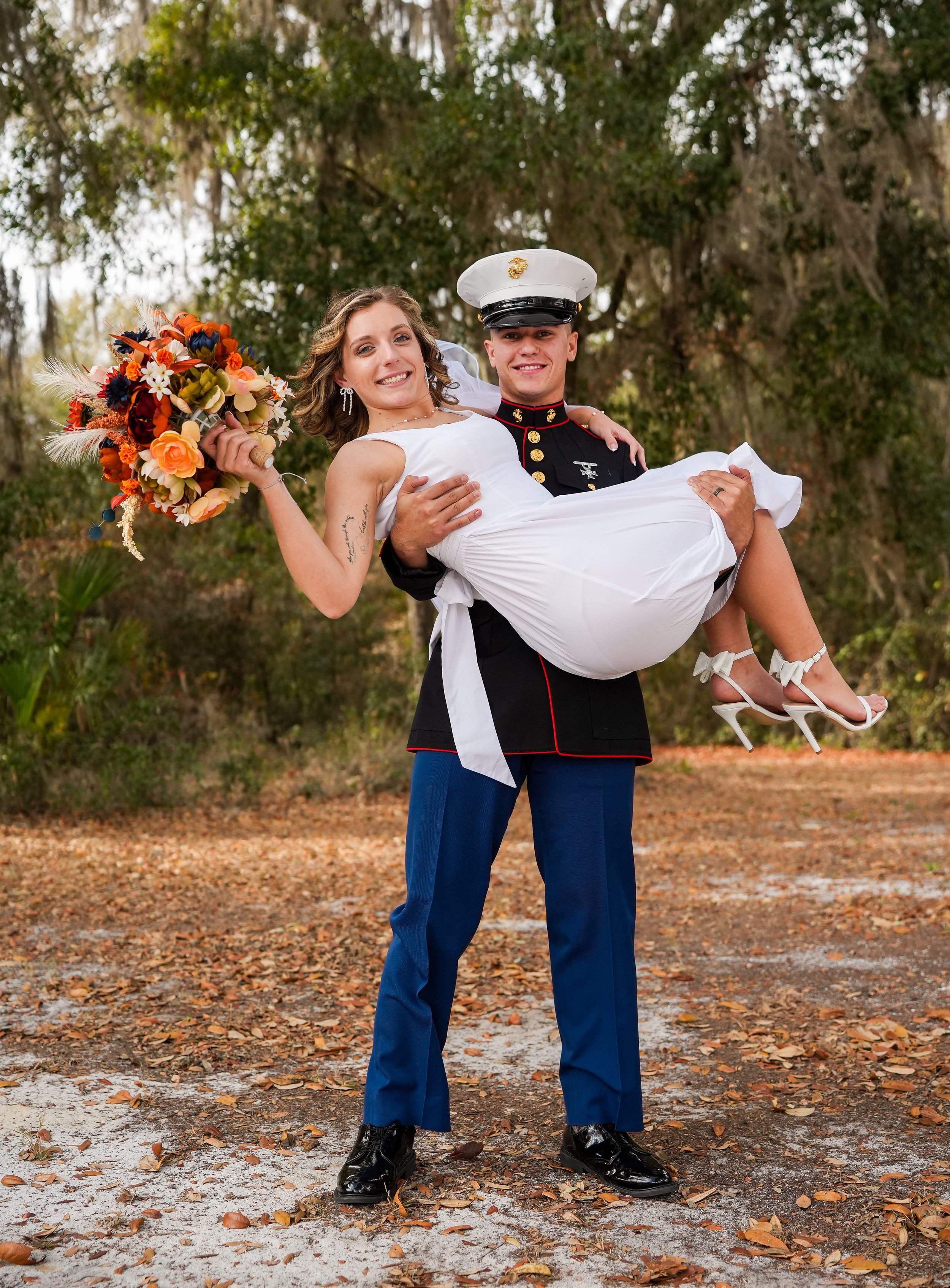 A groom in a United States Marine Corps uniform holding a smiling bride in a white dress and high heels in an outdoor setting with trees and fallen leaves.