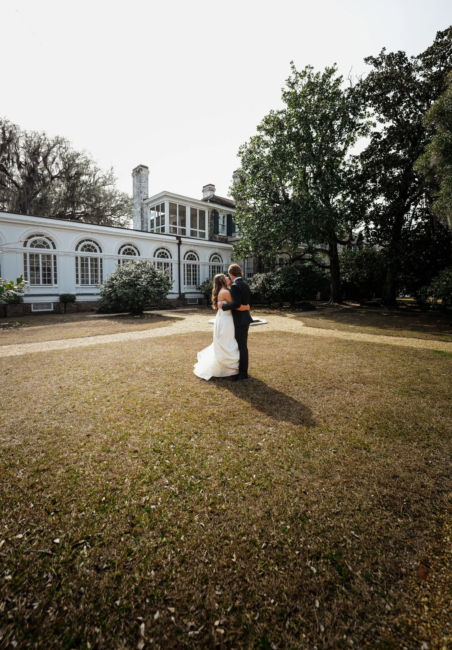 A bride and groom sharing a kiss outdoors in front of a historic house or mansion with large windows and chimneys, surrounded by trees and shrubs.