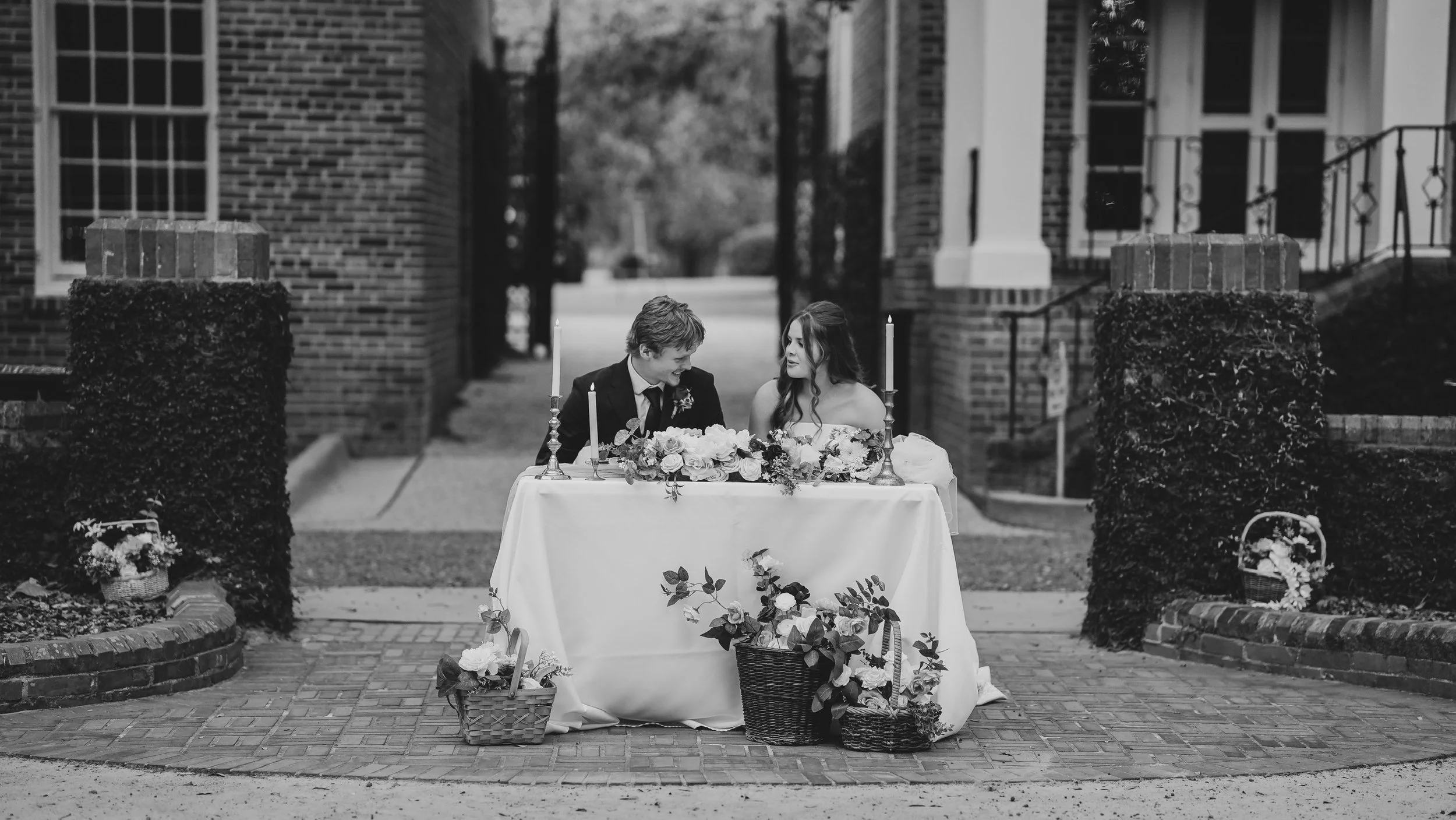 A black and white photo of a wedding reception setting outdoors, with a table decorated with flowers, candles, and baskets full of flowers. There is a bride and groom sitting at the table, smiling and talking to each other.