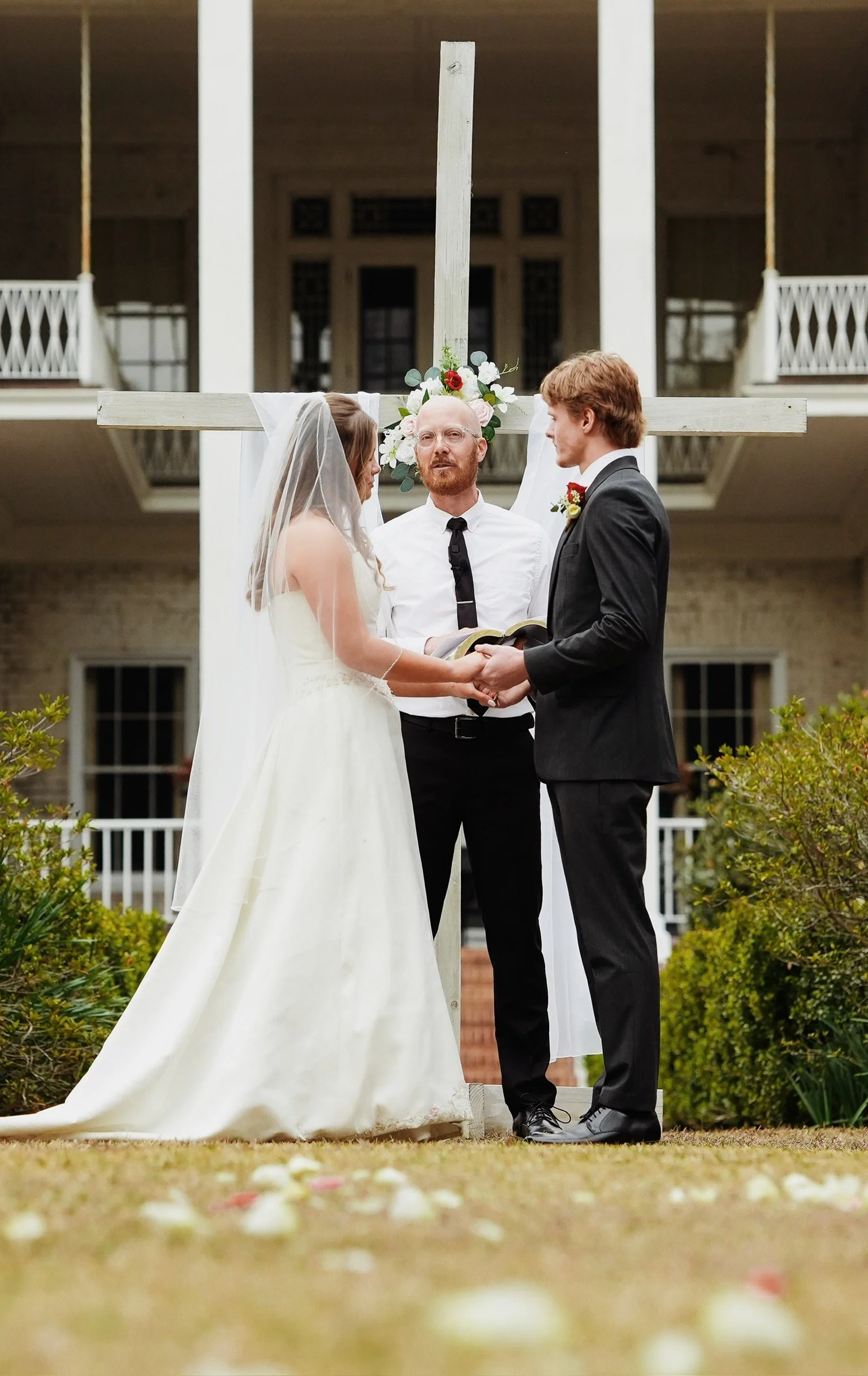 A wedding ceremony with a bride and groom holding hands in front of an officiant, standing under a large cross decorated with flowers outside a building, with flower petals on the ground.