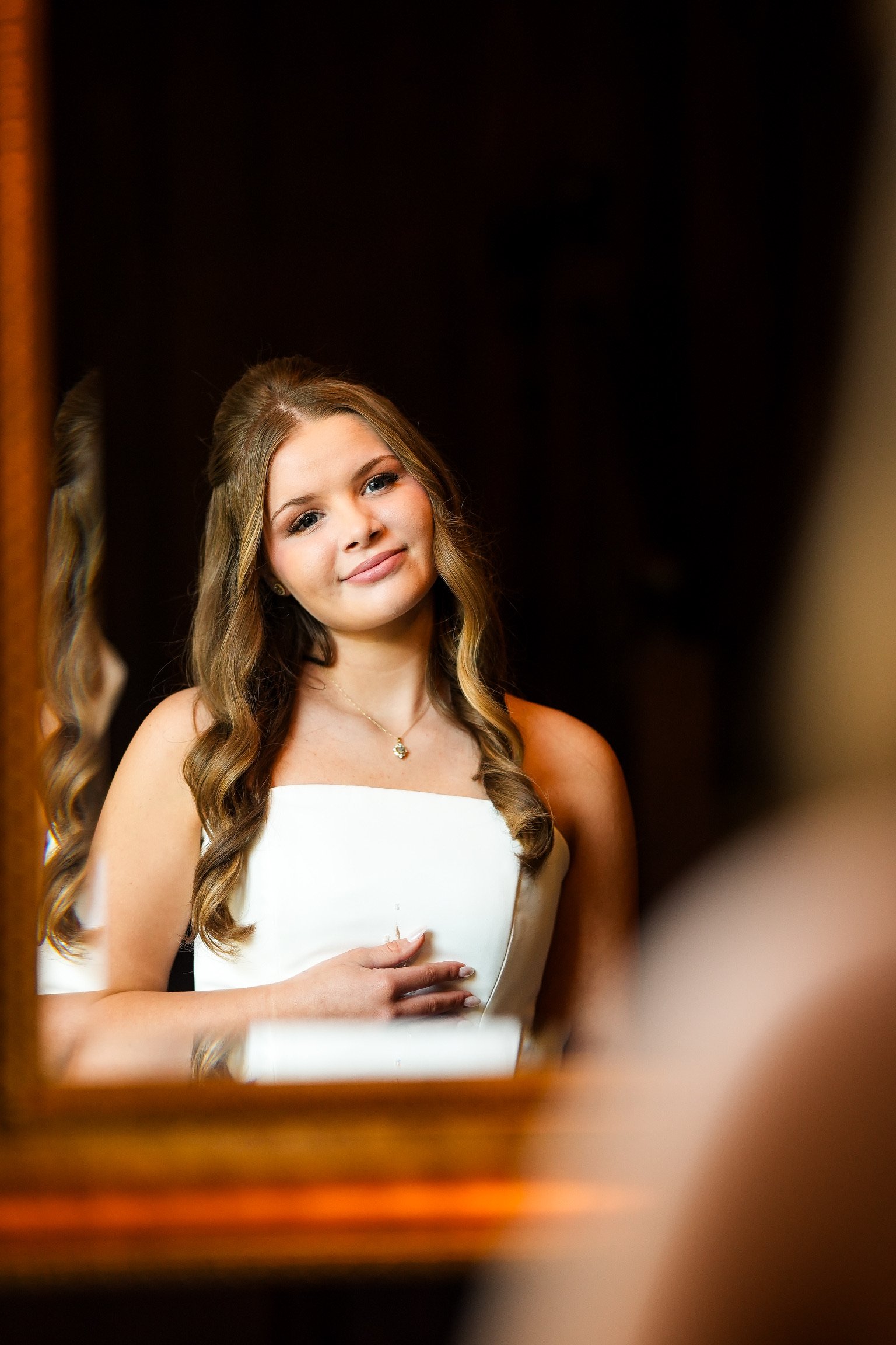 A young woman with long, wavy hair, wearing a strapless white dress and a necklace, is taking a selfie in front of a mirror. She is smiling softly and holding one hand over her stomach. The background is dark, and a portion of a mirror frame is visib