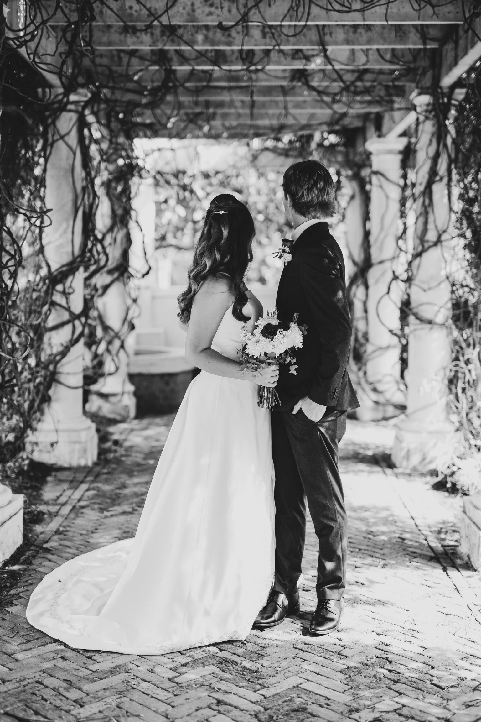 A black and white photo of a bride and groom standing together under a decorative archway made of twisted vines and branches. The bride is holding a bouquet, and the groom is facing her, both dressed in formal wedding attire.