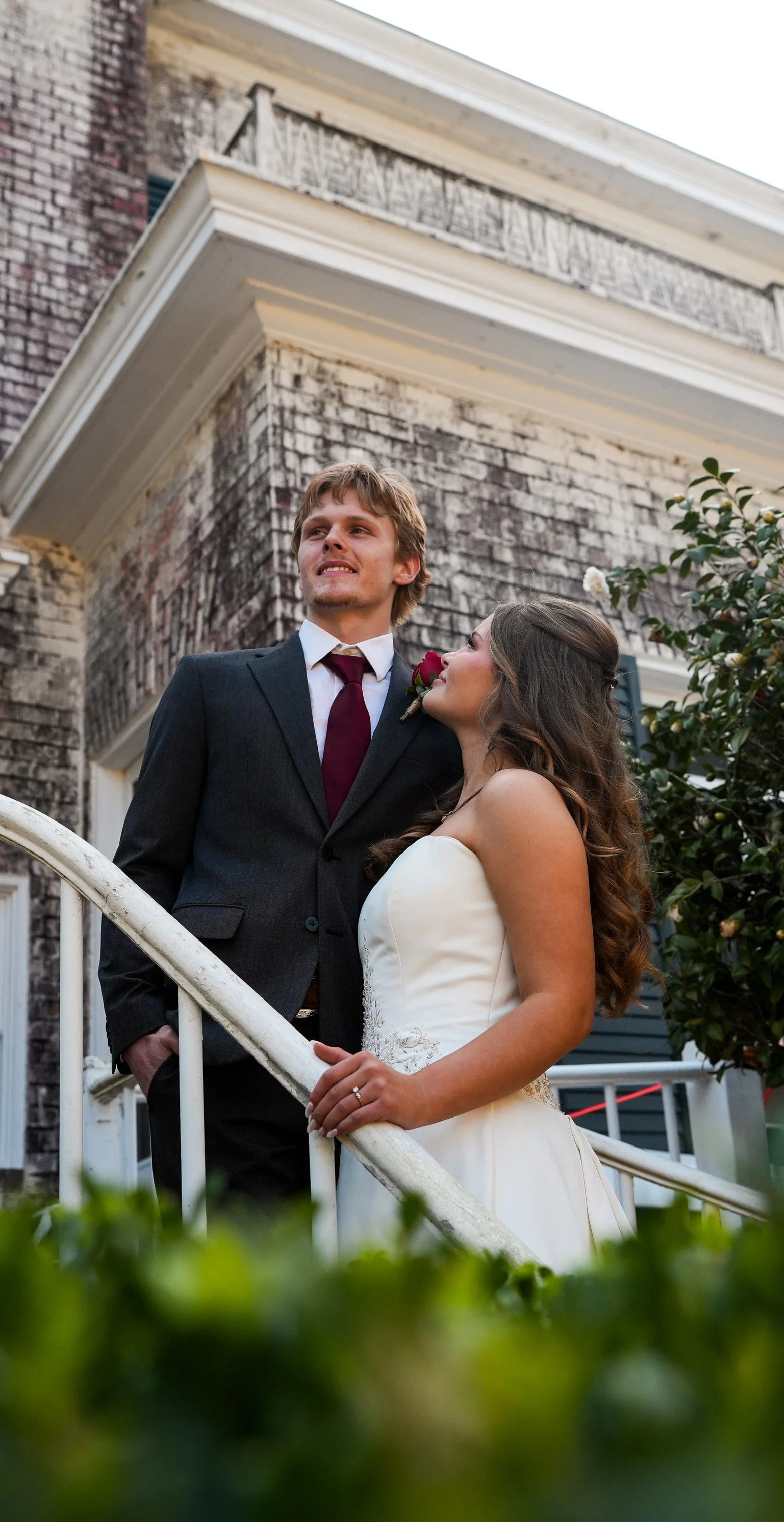A newlywed couple standing on the steps of a brick house, with the groom dressed in a black suit, white shirt, and burgundy tie, and the bride in a strapless white wedding gown, looking at each other affectionately.