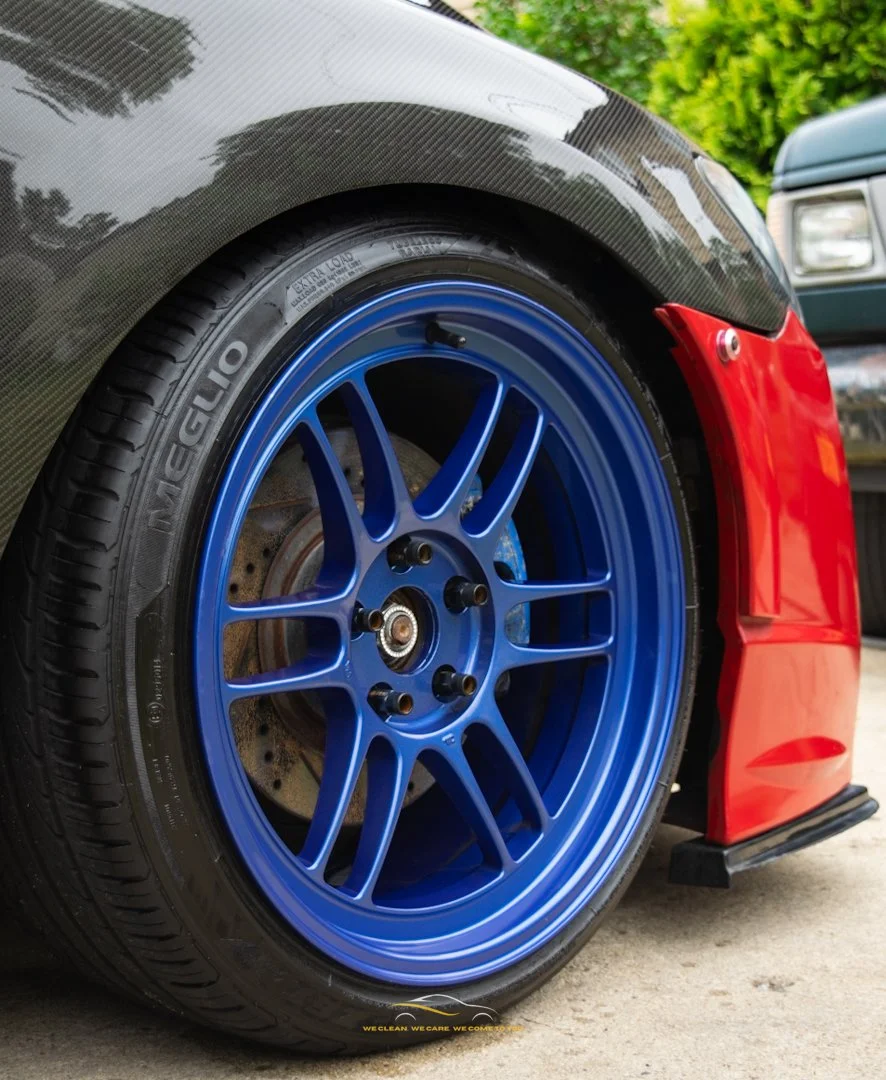 Close-up of a racing car's front wheel with a blue rim, black tire, red front bumper, and a section of a carbon fiber fender, parked outdoors with greenery in the background.