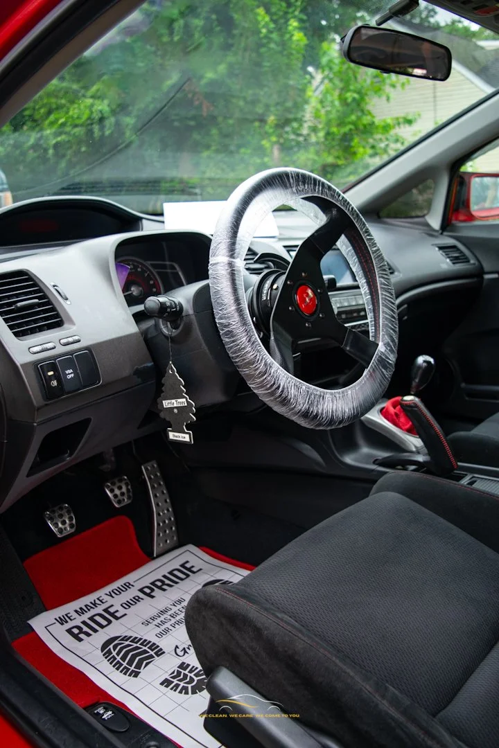The interior of a car showing a steering wheel with a plastic cover, a gear shift, and a black seat. There is a red and black floor mat that says 'We make your ride our pride' and a tree-shaped air freshener hanging from the dashboard. The windshield