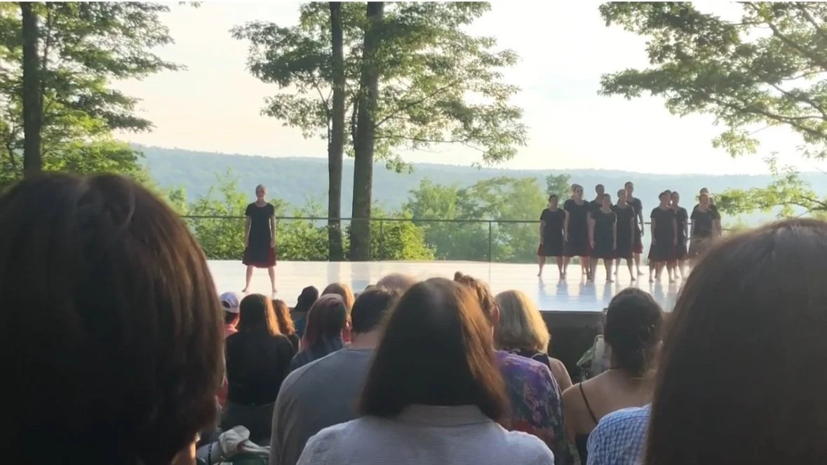 A group of women performing a dance on an outdoor stage with a scenic backdrop of trees and hills. Audience watching from below.