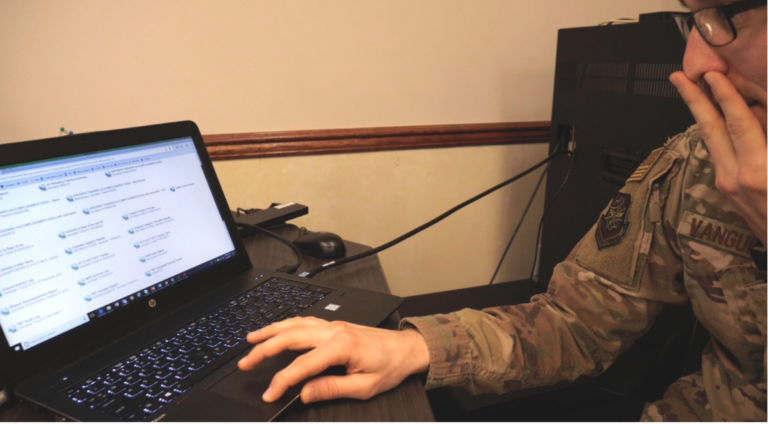 A person in military uniform working on a laptop with a social media page open, sitting at a desk with a monitor, mouse, and keyboard.