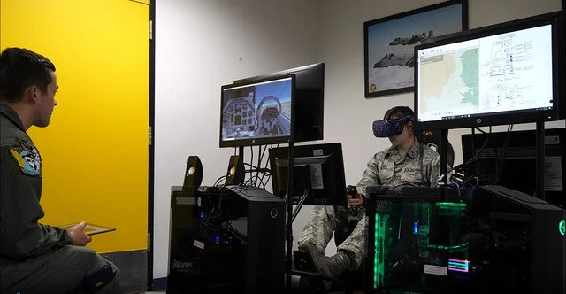Military personnel in a training or simulation room with computers and VR equipment, one person wearing a VR headset and another observing.