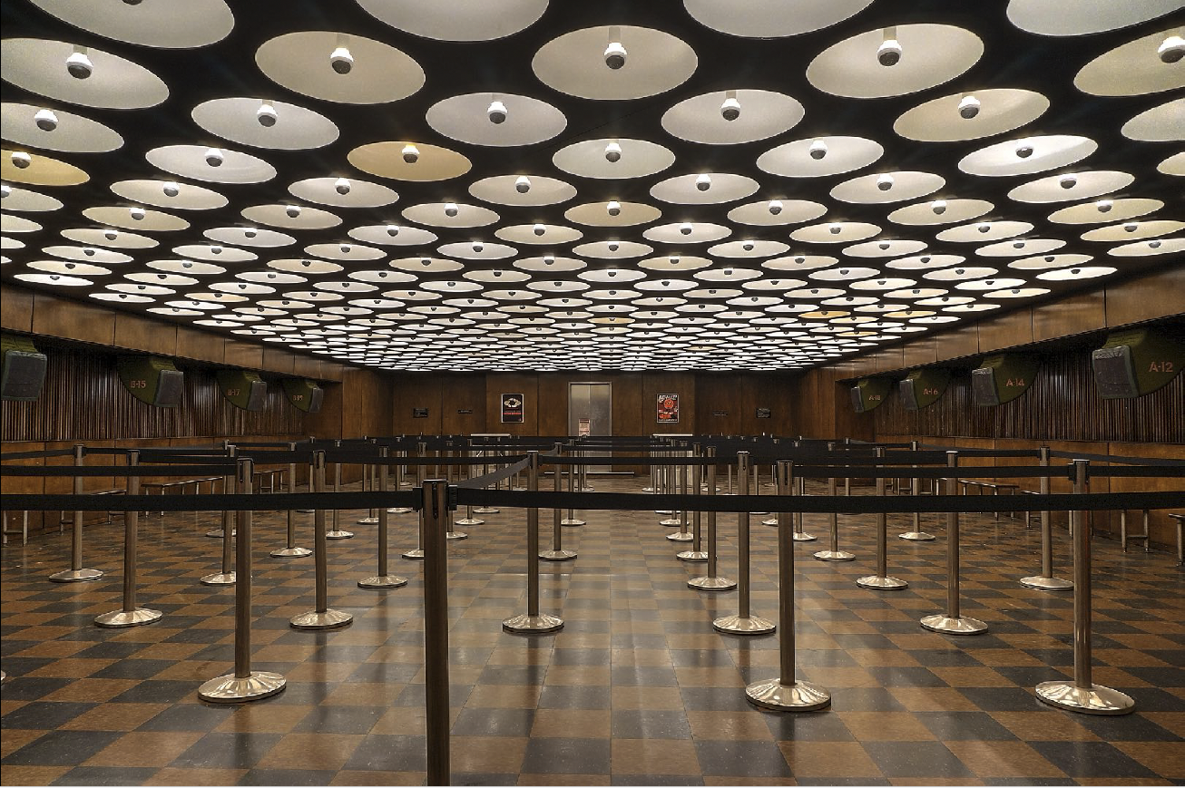 TVA  Miss Minutes with black stanchions and queue barriers, wood-paneled walls, check-in booths labeled with numbers, and a ceiling featuring a pattern of circular lights.