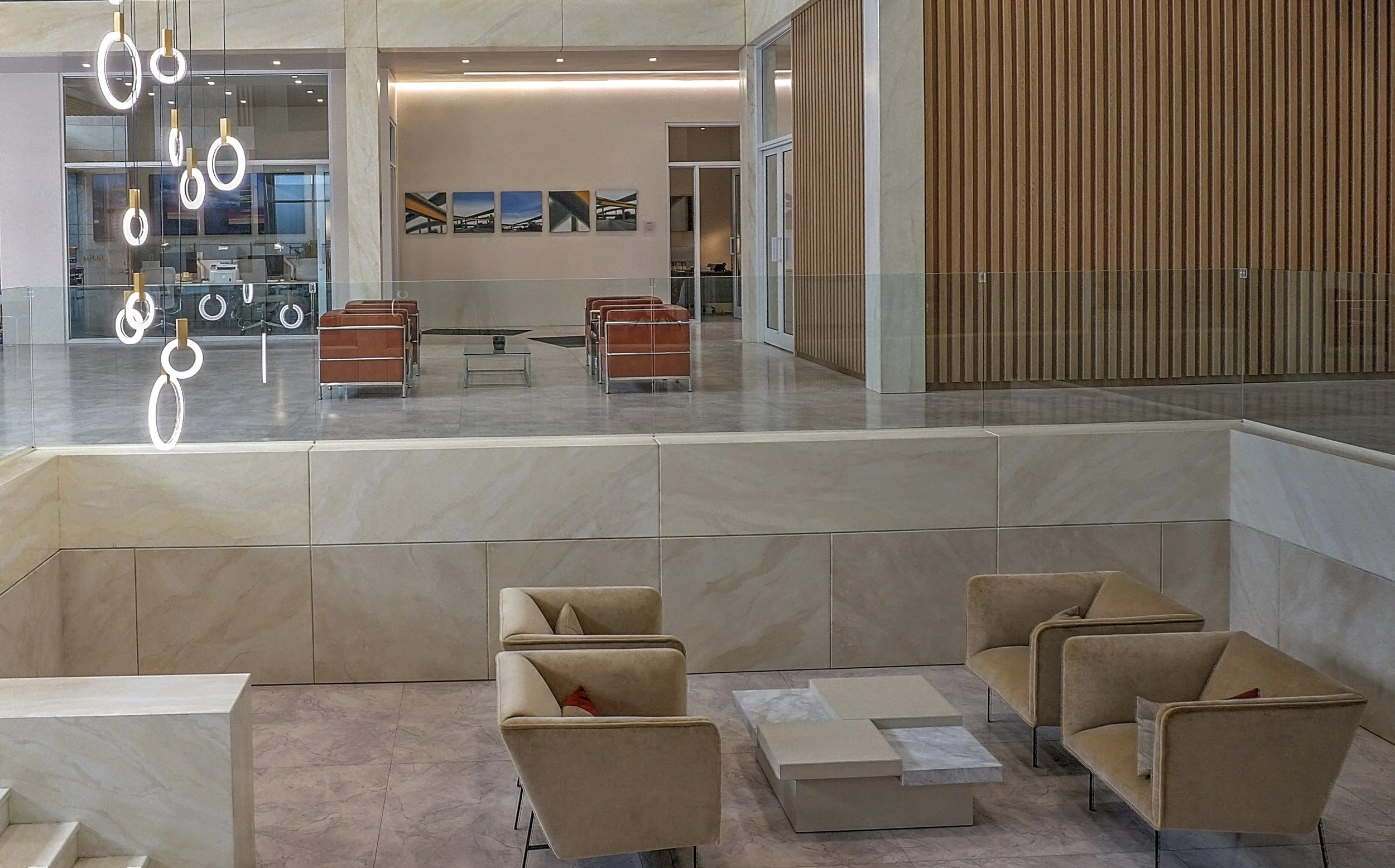 She Hulk Law Modern office lobby with beige seating area, glass railing, and wooden wall paneling, illuminated pendant lights, and artwork on the distant wall.