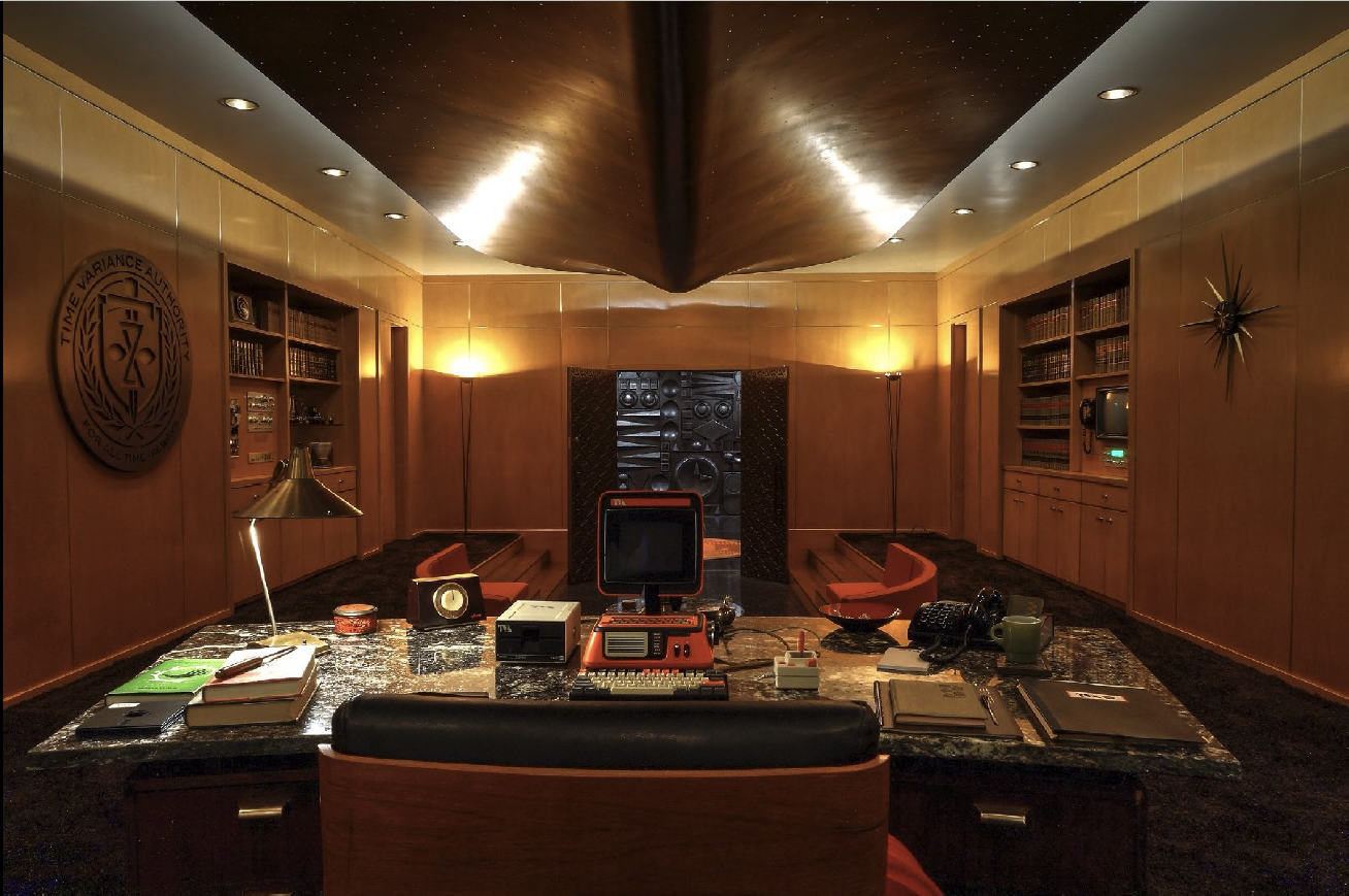 Mid-century modern office with wood-paneled walls, built-in bookshelves, and a central desk cluttered with office supplies, books, and electronics. There is a large wall clock, a computer, and vintage decor including a sculpture and a stylized starbu