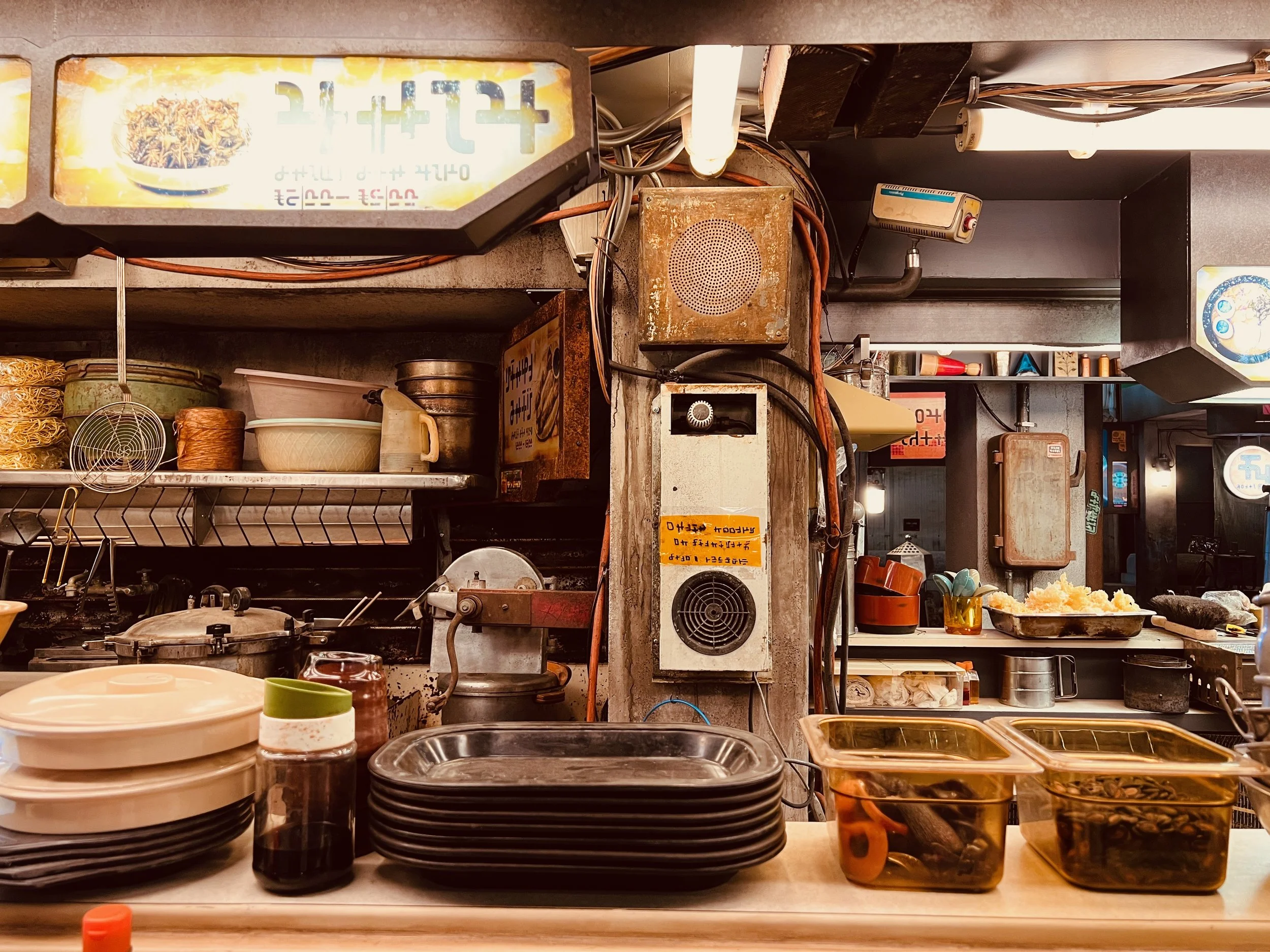 Rebel Moon Daggas noodle bar restaurant kitchen with stacks of plates, jars, containers filled with food, and kitchen equipment like a rice cooker and pots.