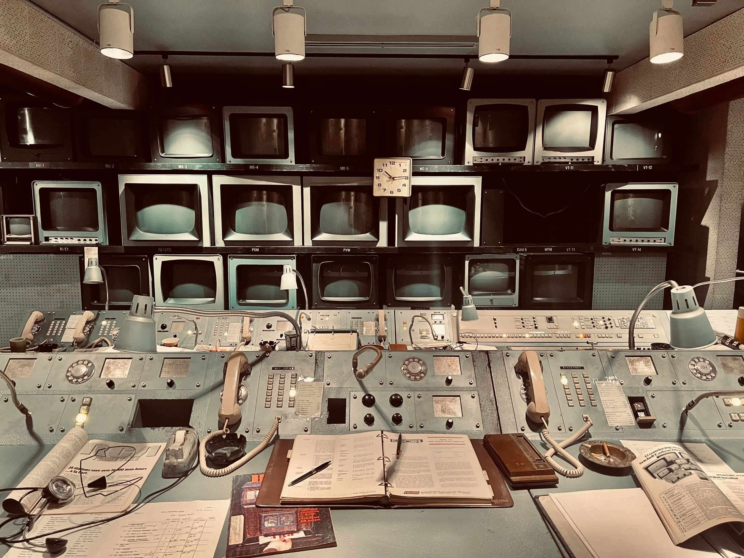 Saturday Night movie broadcasting 1975 Television production control room with multiple monitors and equipment, vintage style, with papers, books, and telephones on the desk.