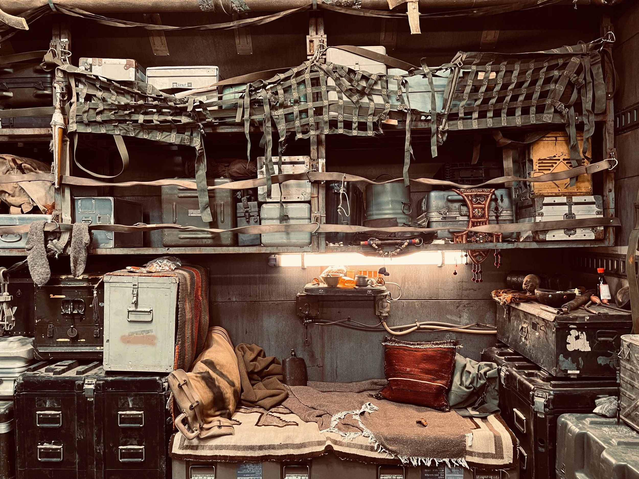 Interior of Kai's Spaceship with shelves filled with vintage metal containers, tools, and equipment. The lower area has a bed with a blanket, bag, and small items, surrounded by black storage cases and a pillow.  Rebel Moon
