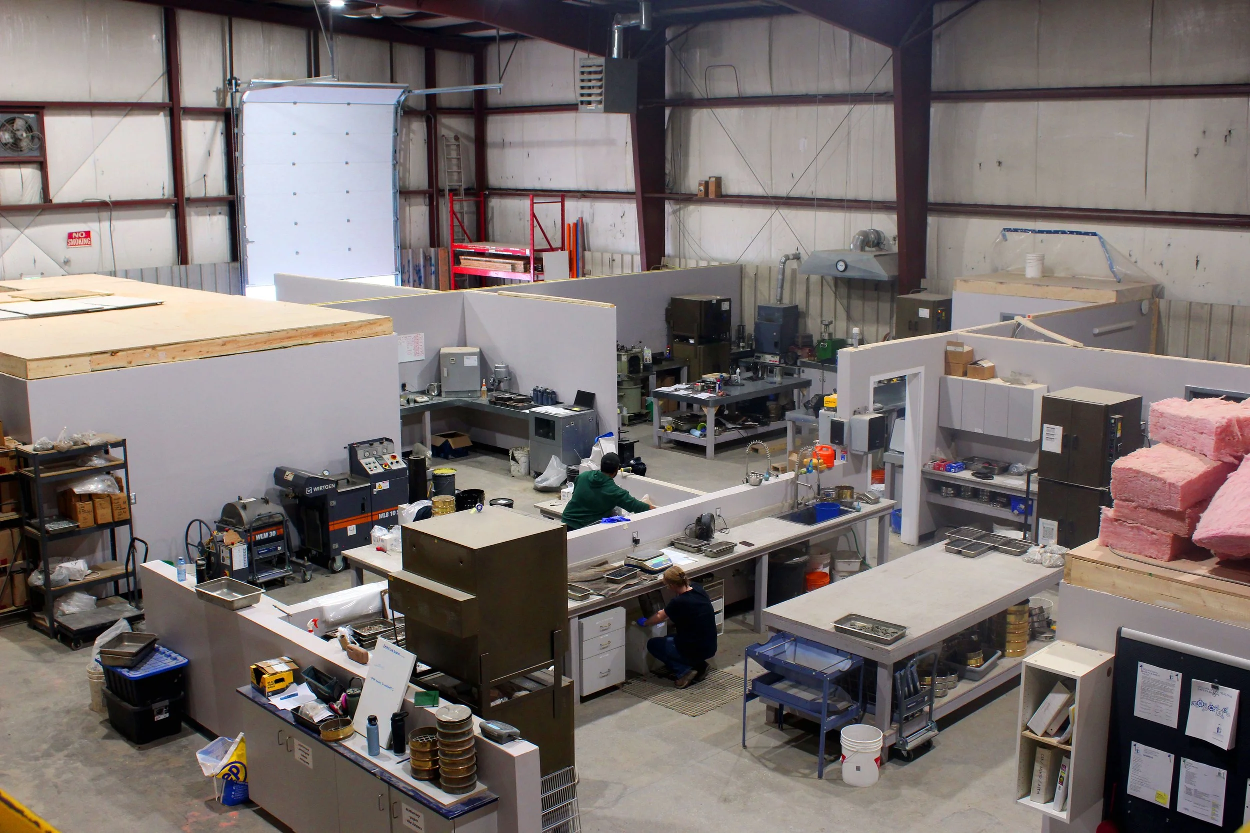 Mezzanine view of different workstations in the Harbourside Geotechnical and Materials Laboratory. A large bay door is half-open in the background, and two individuals can be seen at different workstations.