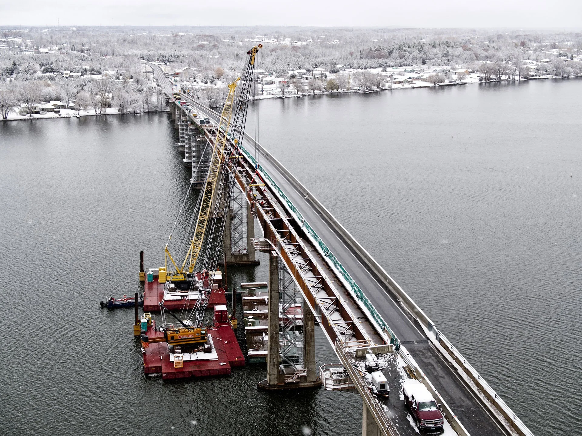 An aerial view of the Bay of Quinte Skyway Bridge under construction. Two massive cranes are supported by barges beneath the bridge. One lane remains open to traffic during construction. A light dusting of snow covers sections of the bridge. Ontario.