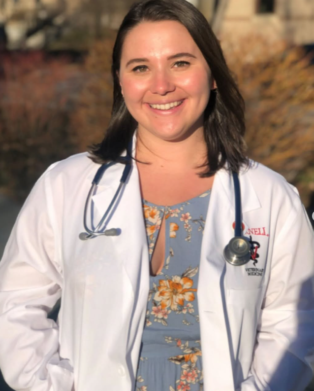 A woman smiling outdoors wearing a white medical coat with a stethoscope around her neck.