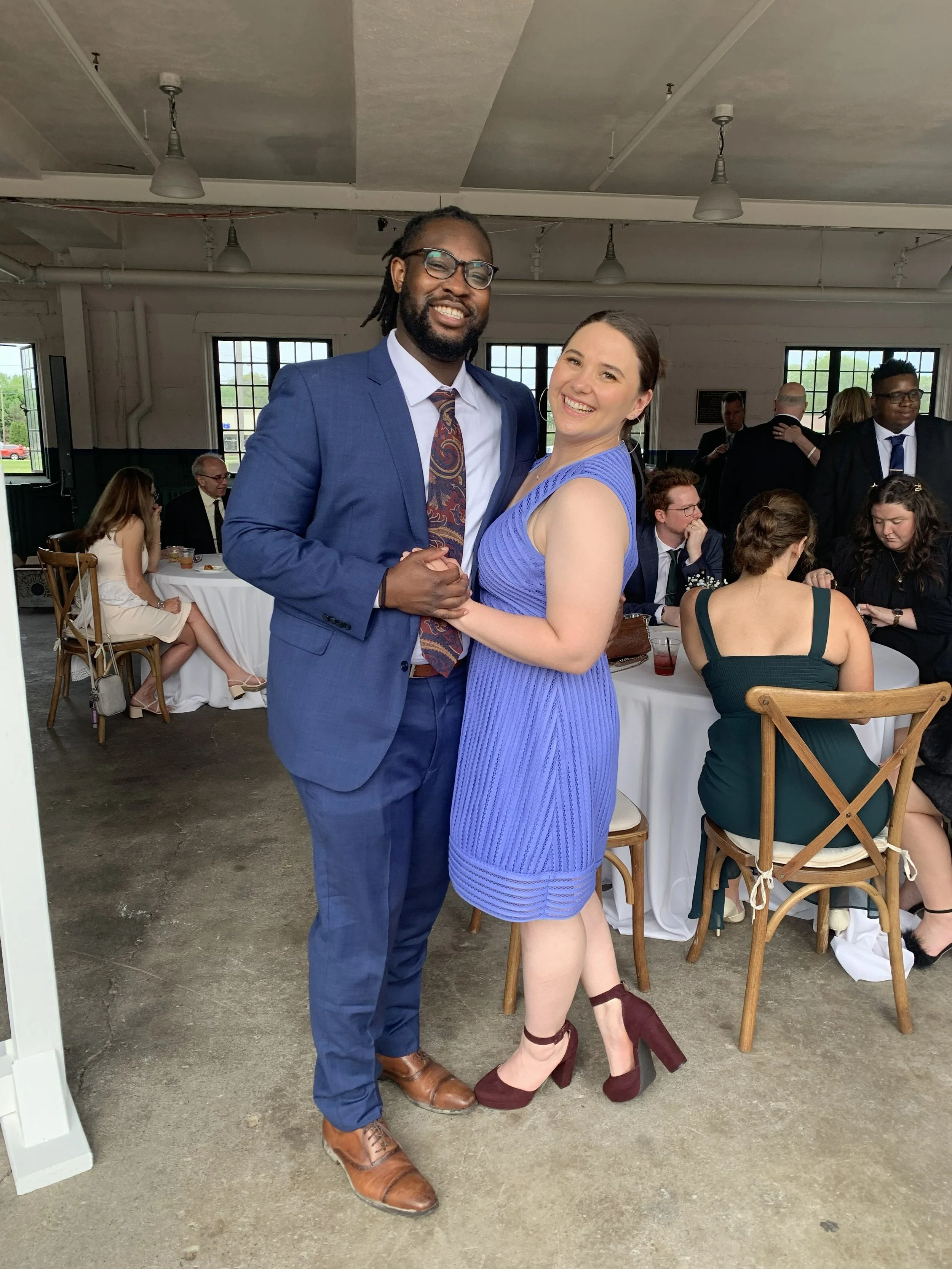A smiling couple dancing at a wedding reception indoors, with guests seated at tables in the background.