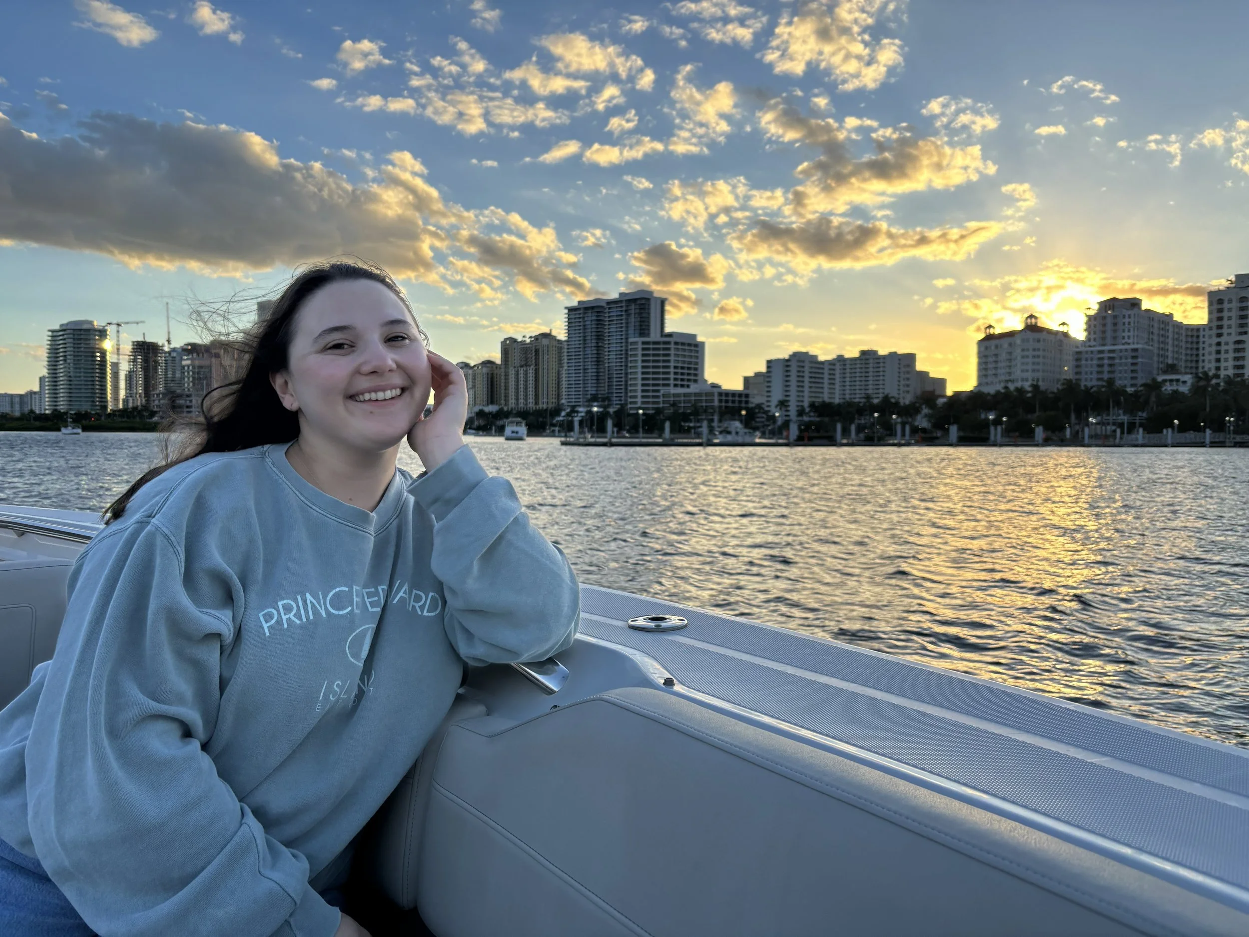 Smiling woman with long hair in a gray Prince Edward Island sweatshirt sitting on a boat, with a city skyline and sunset in the background across the water.