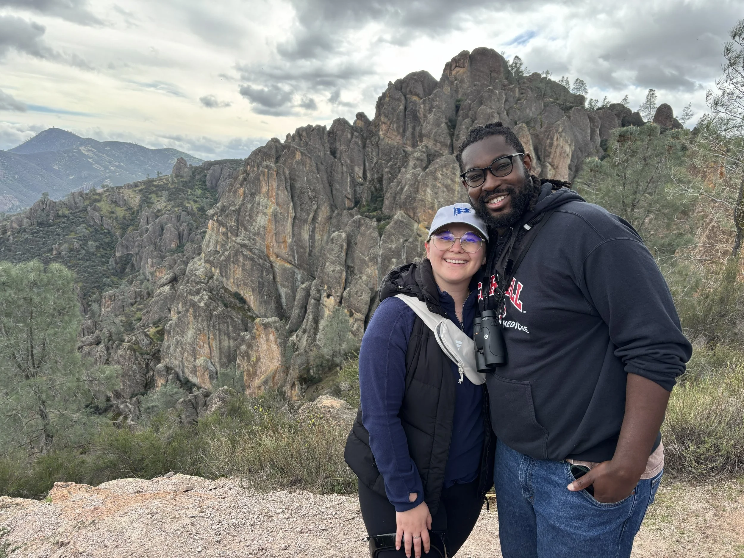 A smiling couple stands close together outdoors with mountains and rocky cliffs in the background. The woman wears glasses, a baseball cap, and a vest, while the man wears glasses, a black hoodie, and has binoculars around his neck.