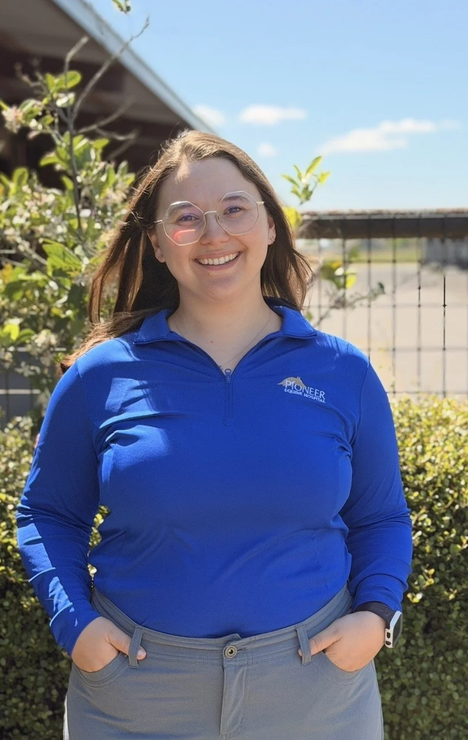 A woman smiling outdoors in front of a hedge, wearing glasses, a blue quarter-zip shirt with 'Pioneer Equine Hospital' logo, and gray pants.